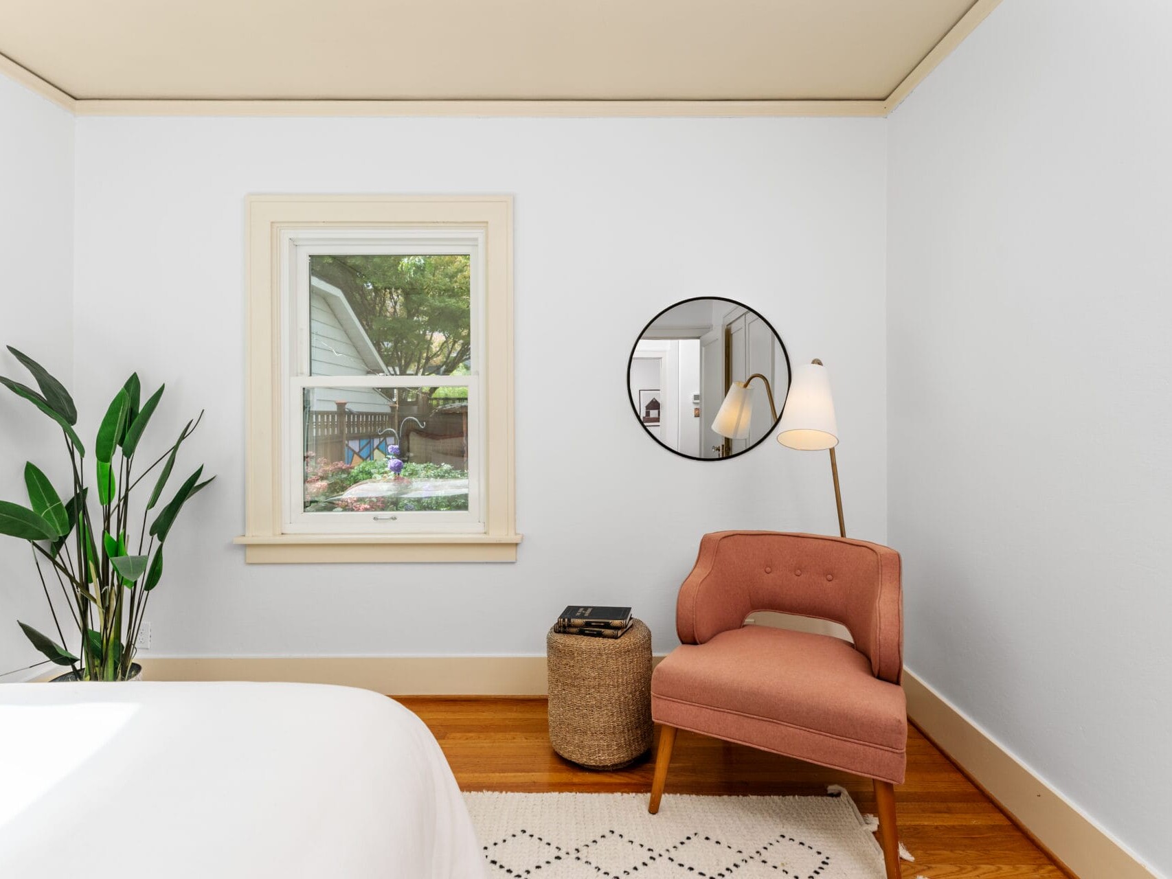 A cozy bedroom corner with a round mirror on the wall, a pink upholstered chair, a small wooden side table with books, and a modern floor lamp. A large green plant sits by a window with a view of greenery outside. The floor is wood with a white rug.