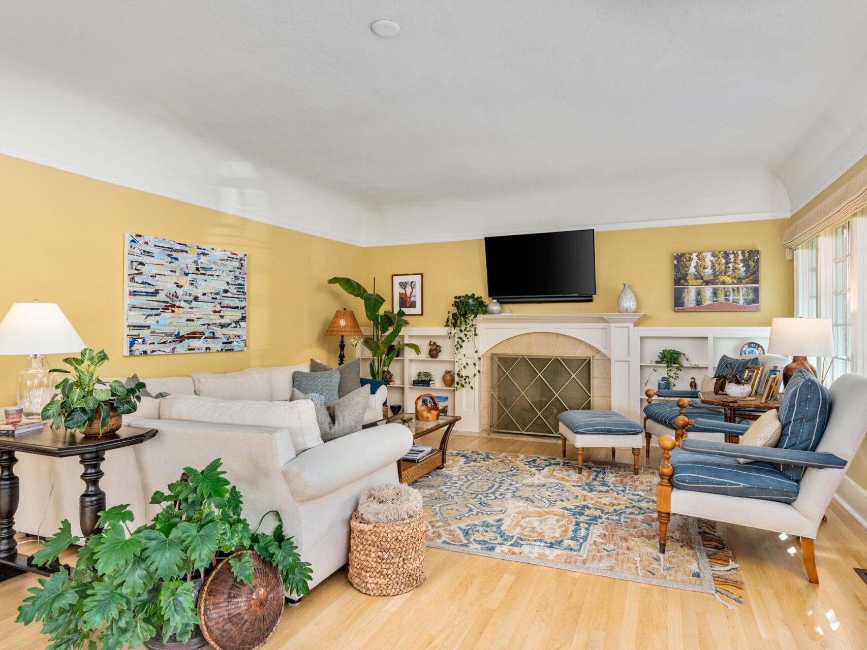 A cozy living room with a white sofa, green plants, and an abstract painting on a yellow wall. A fireplace with a TV above it is flanked by armchairs. Natural light streams through a window on the right, illuminating a patterned rug on the wooden floor.