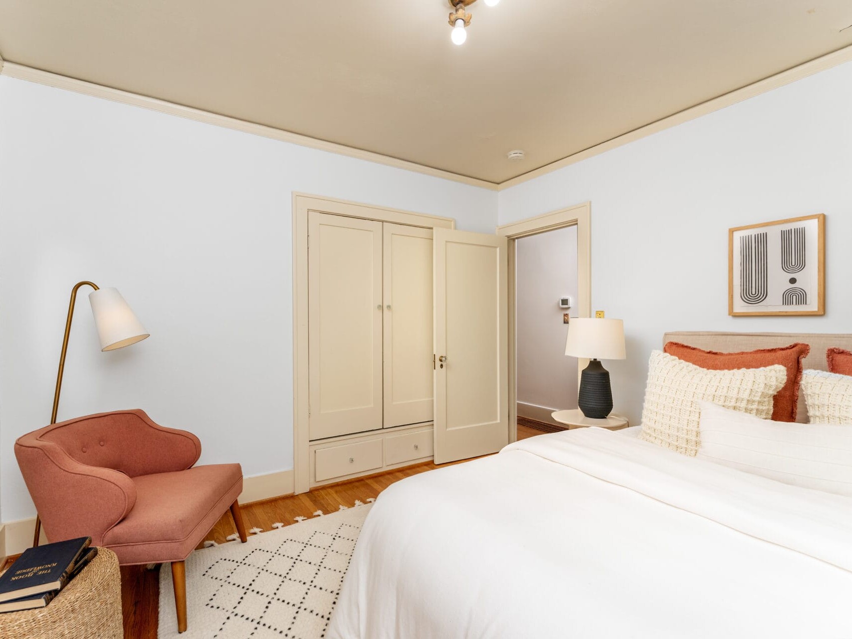 A cozy bedroom with a neatly made bed featuring white linens and textured pillows. A pink armchair and a standing lamp are in the corner. A wardrobe with closed doors is on the wall next to three abstract framed artworks. A small rug covers the floor.