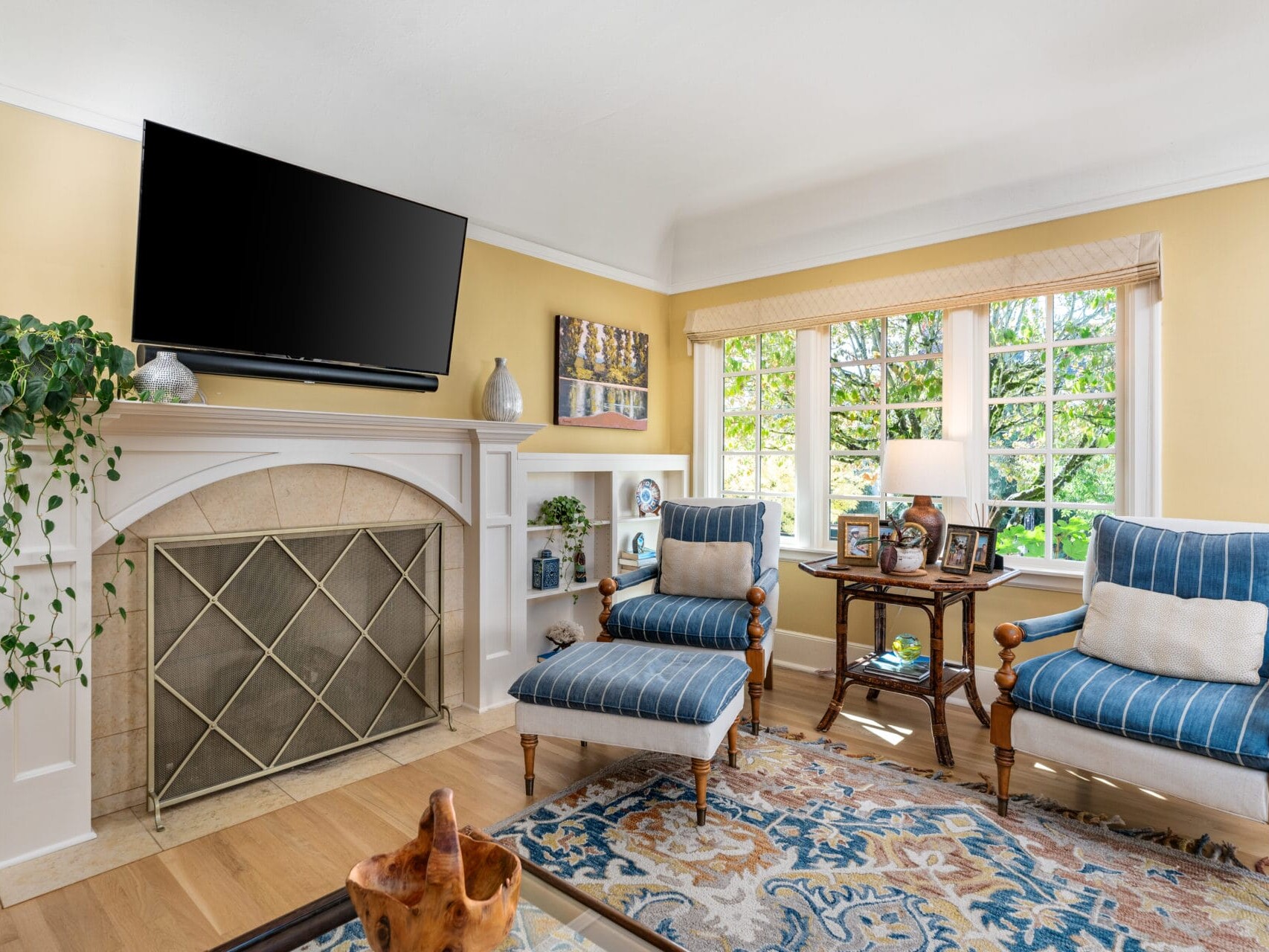 A cozy living room with yellow walls featuring a fireplace and a large TV above it. Two blue striped chairs with white cushions sit next to a window. A coffee table with decorations and a patterned rug complete the inviting space.