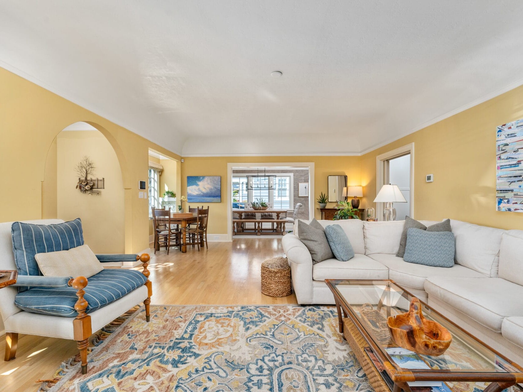 A spacious living room with a cream-colored sectional sofa and striped armchair on a patterned rug. Bright walls, arched doorway, and a dining area in the background. Decor includes a nautical-themed painting and a wooden coffee table.