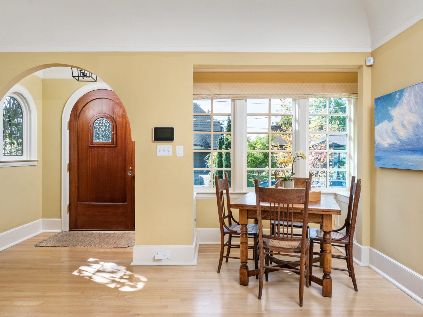 A cozy dining room with light yellow walls features a wooden table and four chairs. Large windows bring in natural light. An arched wooden door is to the left, and a painting hangs on the wall to the right. A small rug is placed by the entrance.