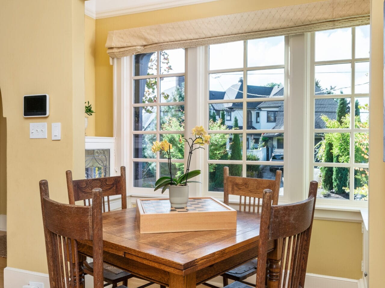 A cozy dining nook with a wooden table and four chairs is situated by a large window with a view of a neighborhood. Sunlight illuminates the space, and a potted plant decorates the table. Walls are painted a warm yellow.