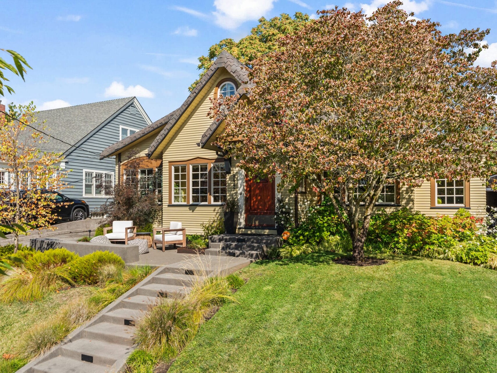 A charming house with a steep roof, surrounded by a lush green lawn and a large tree. There are two white chairs on a paved walkway leading to the entrance. A neighboring house and a parked car are visible in the background.