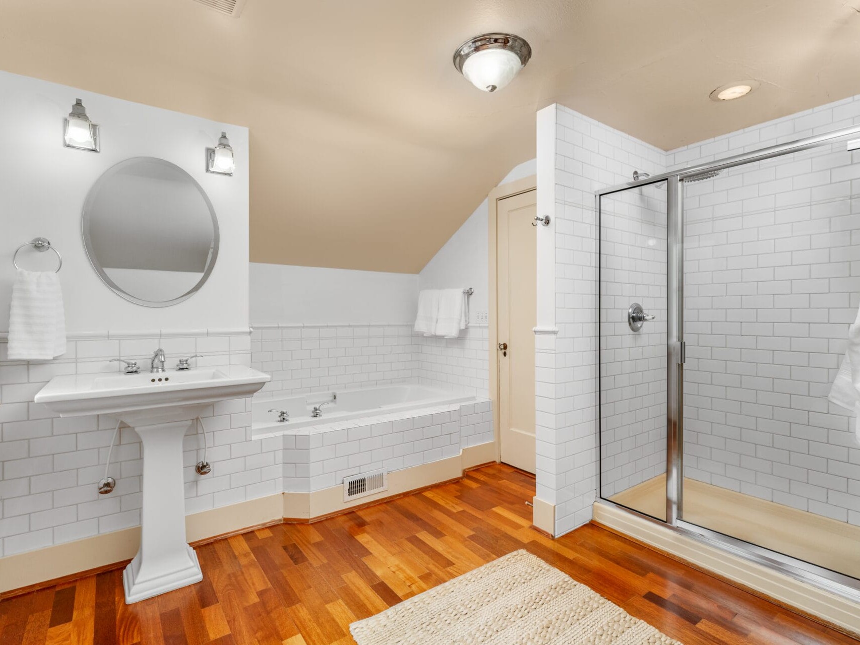 Bathroom with white tiled walls, a pedestal sink, round mirror, and bathtub. A glass-enclosed shower is on the right. The floor has warm-toned wood, and a beige rug is on it. Two wall lamps are above the sink.