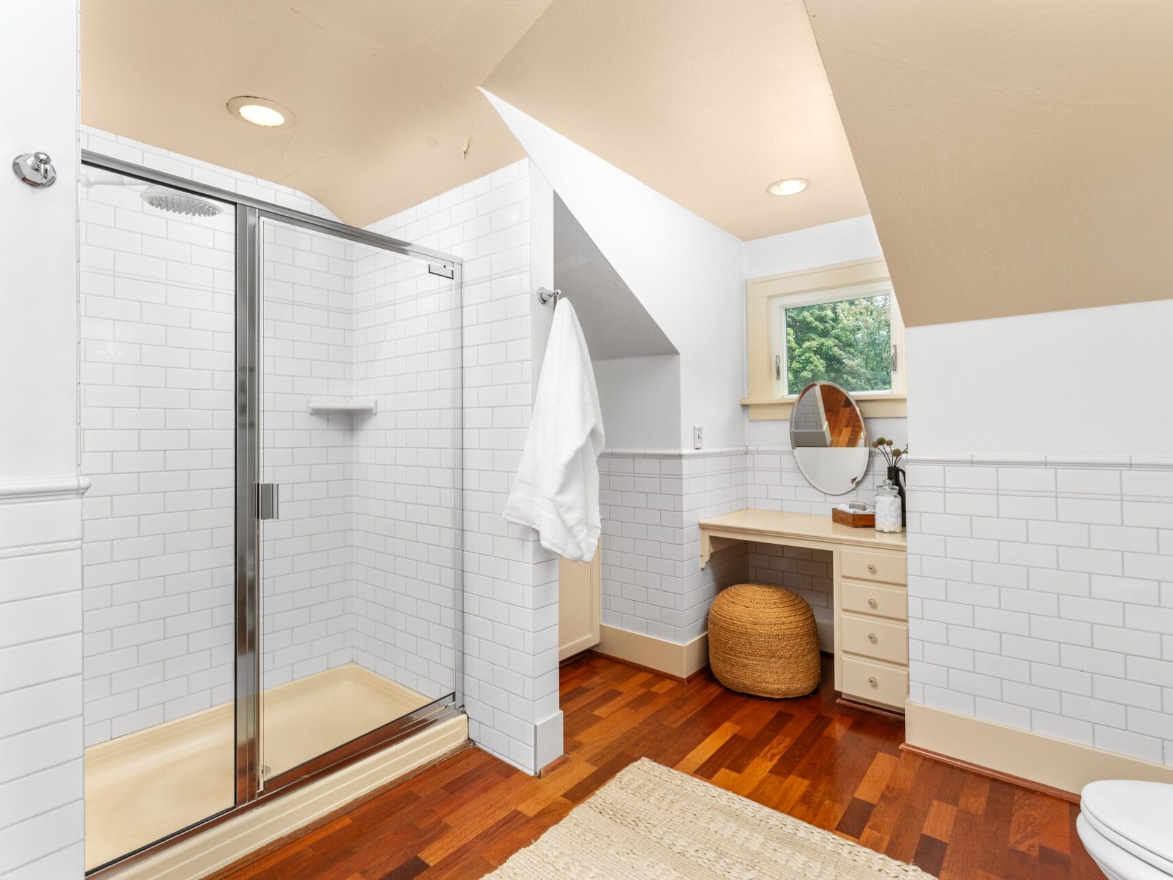 A modern bathroom featuring a glass-enclosed shower with white subway tiles, a wooden floor, a small vanity with a round mirror, and a woven basket under the counter. A towel hangs near the shower, and a window lets in natural light.