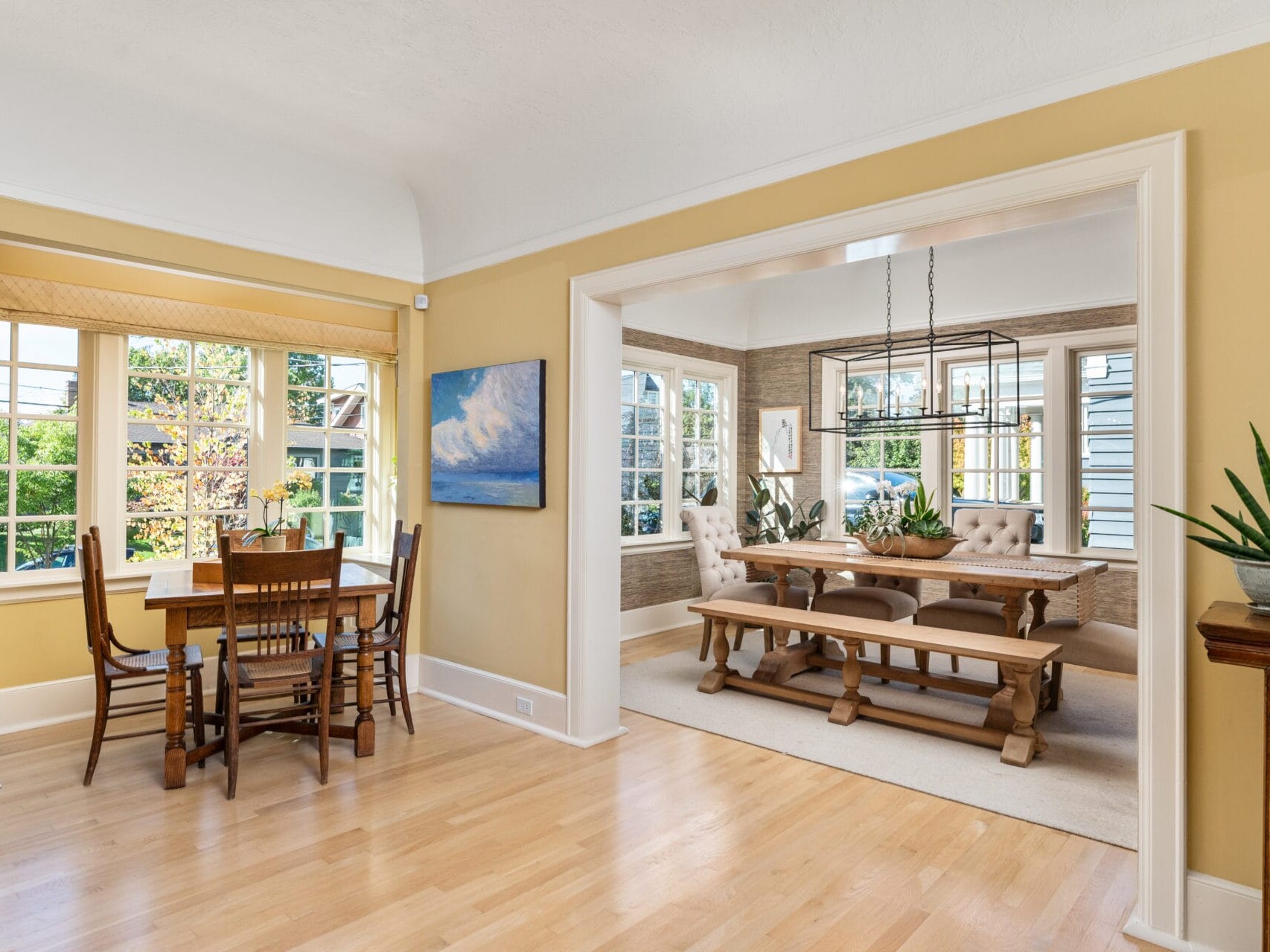 A bright dining area featuring two tables. One sits in a cozy sunlit corner with six wooden chairs by a window. The second table is larger, with cushioned chairs, set under a black chandelier, surrounded by windows and framed by light walls.