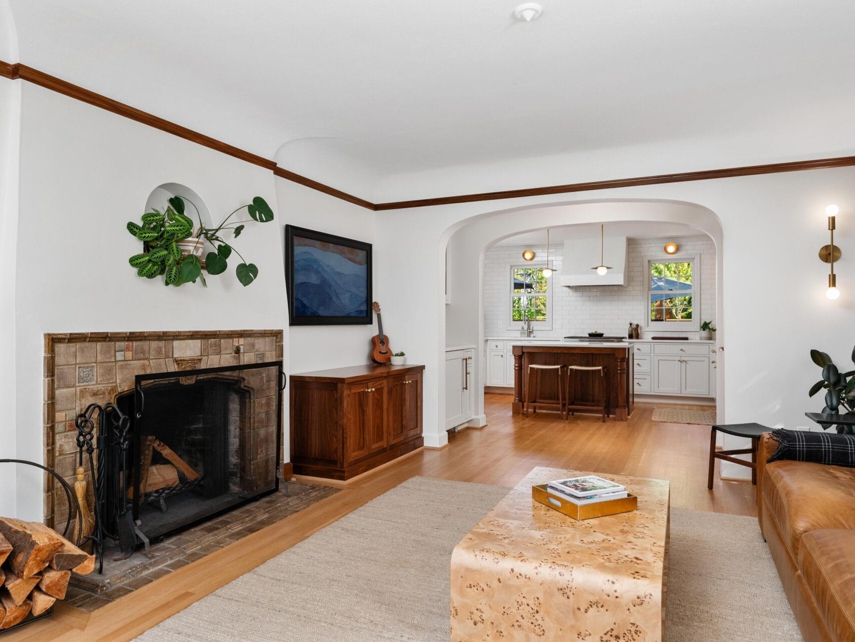 A cozy living room with a fireplace, a plant, and a wall-mounted TV. To the right, a tan leather sofa and a wooden coffee table. Through an archway, a view of a white kitchen with wooden floors and a center island. Natural light brightens the space.
