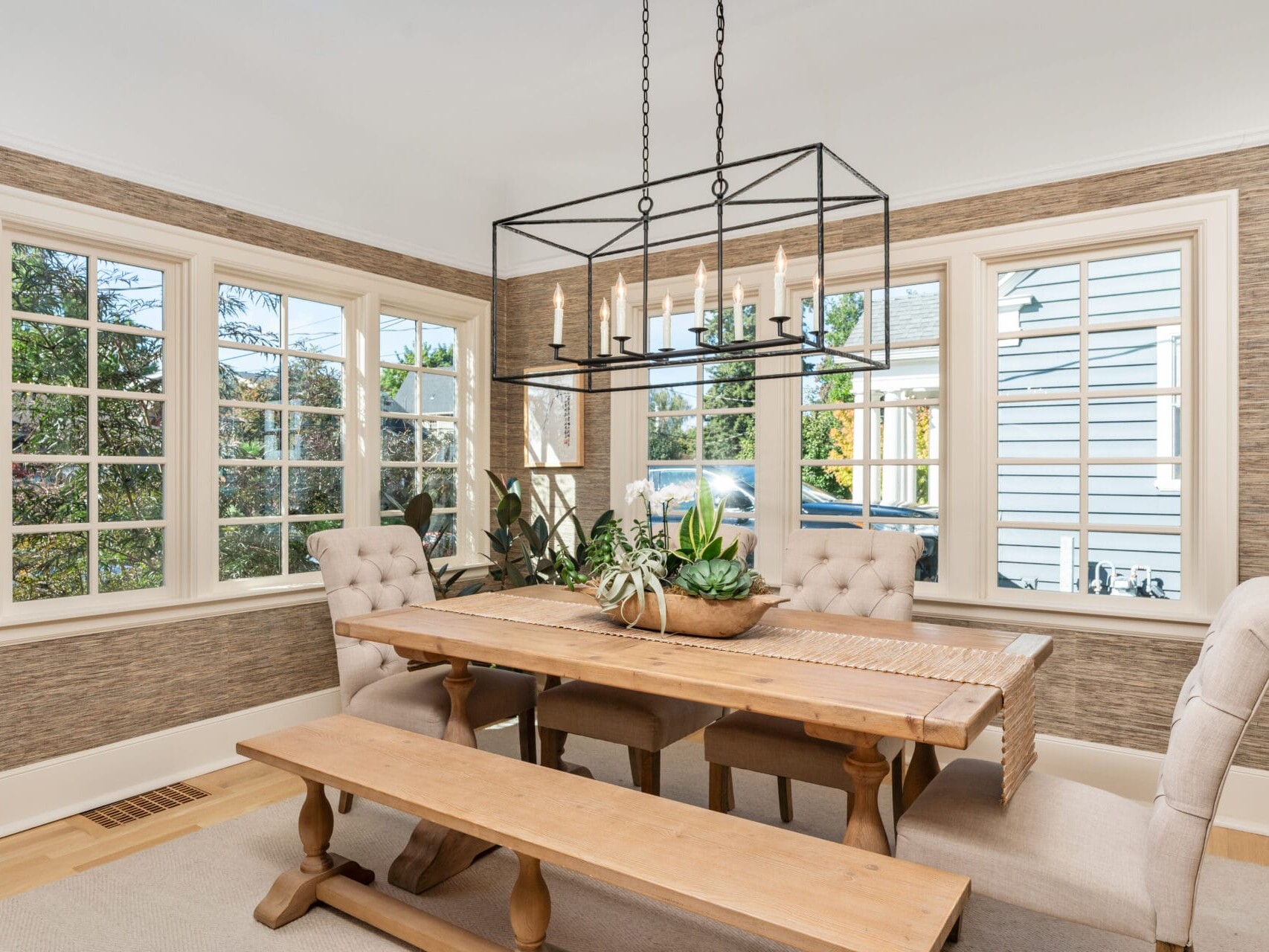 A cozy dining room with a wooden table and benches, surrounded by cushioned chairs. A modern rectangular light fixture hangs above. Large windows provide natural light, and green plants decorate the space. Textured walls add warmth.