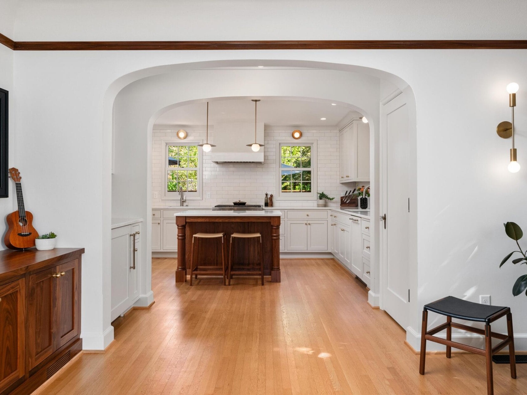 A bright kitchen with white walls and cabinets, wooden floors, and a central island with two stools. The room features two windows, pendant lights, a wall-mounted guitar, potted plants, and a wooden sideboard.