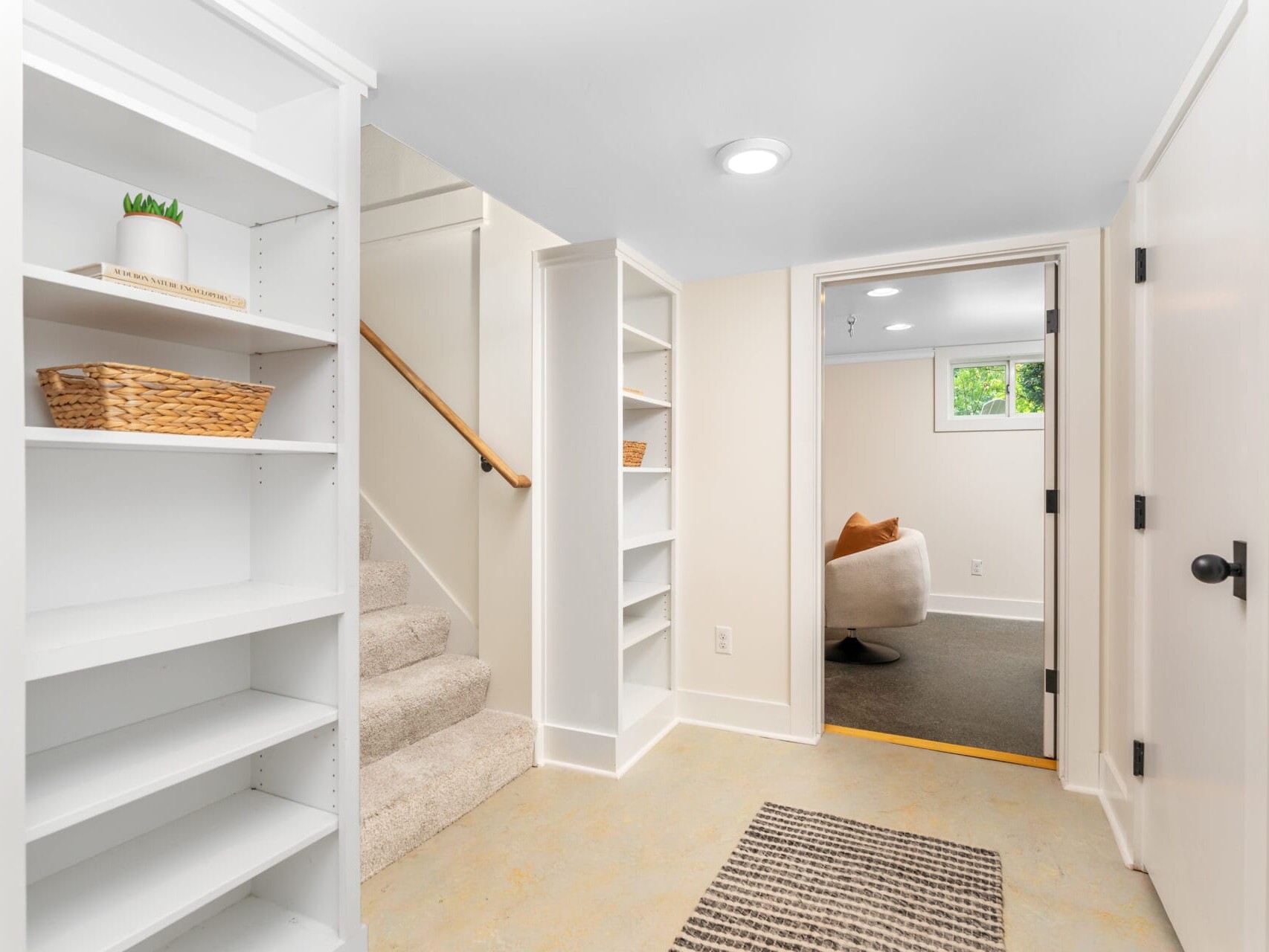 A bright basement hallway with white built-in shelves on the left, featuring wicker baskets and a small plant. Carpeted stairs lead up, and a rug lies on the beige floor. An open door reveals a room with a chair and window in the background.