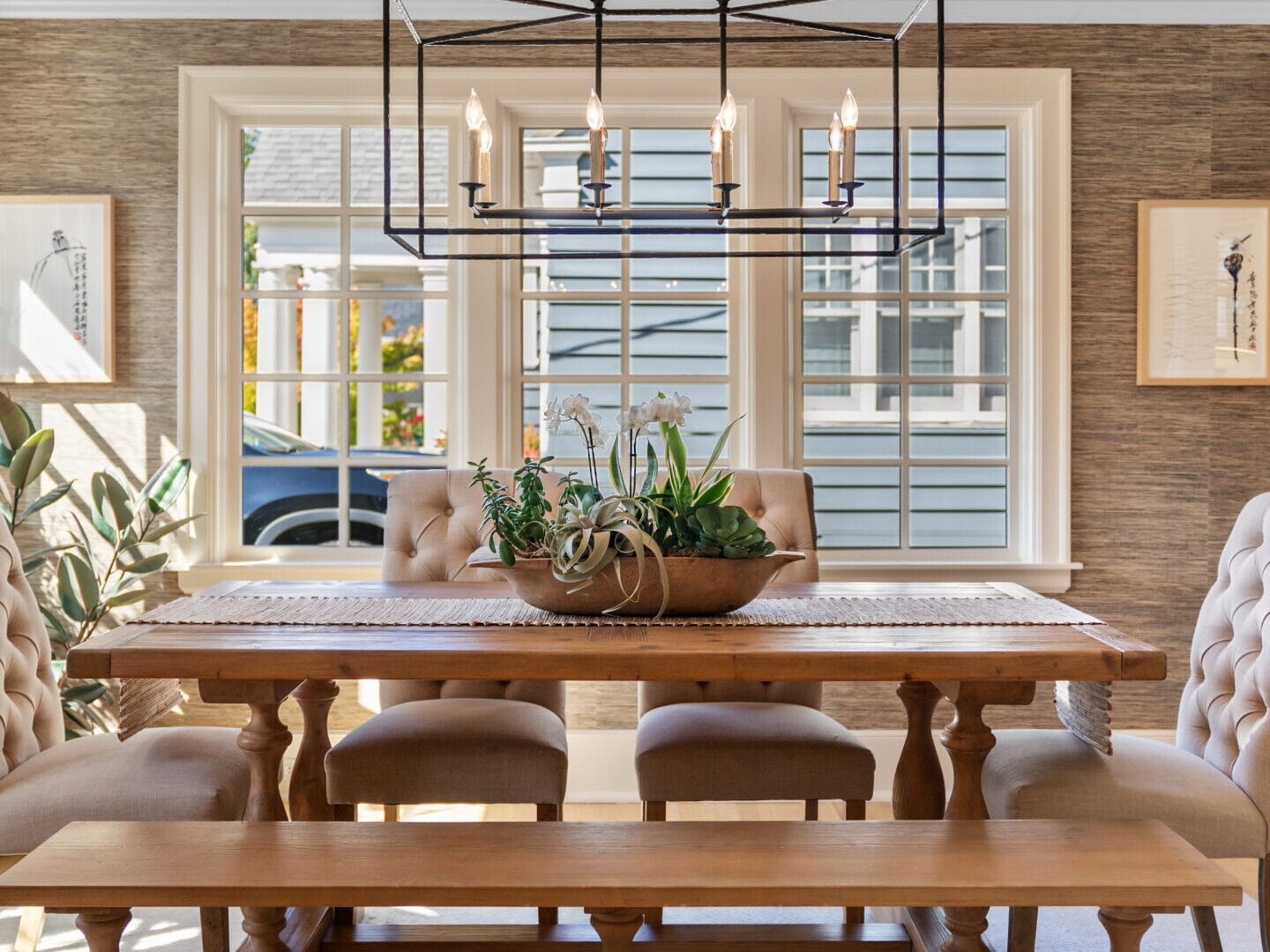 A dining room with a wooden table, cushioned chairs, and a bench. A rectangular black chandelier hangs above. Large windows in the background let in natural light. A decorative plant arrangement sits on the table. Walls are neutral-toned.
