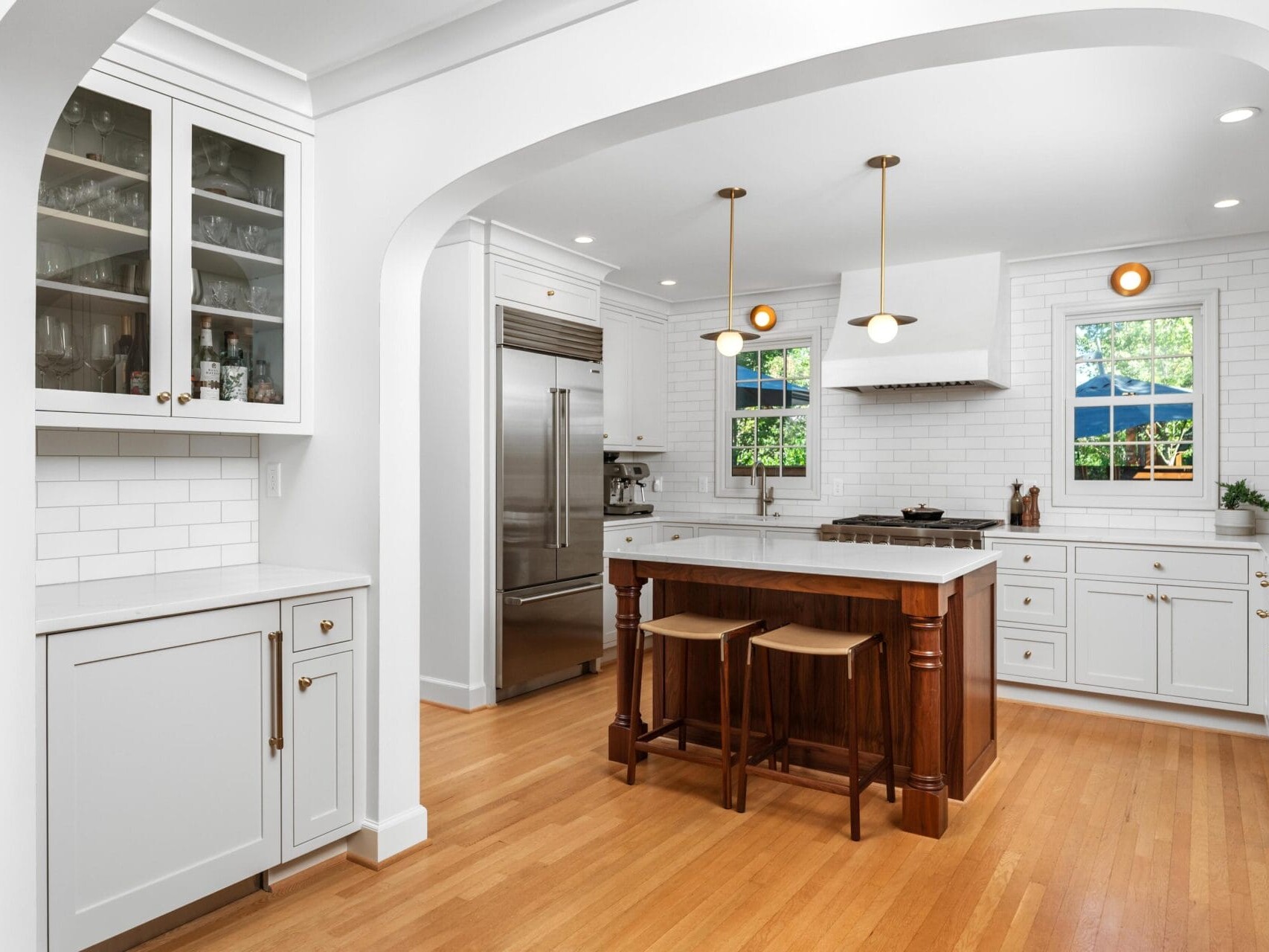 A modern kitchen with white cabinets, a large stainless steel fridge, and a central island with wooden stools. Pendant lights hang above the island. An archway leads into the kitchen, and windows provide natural light.
