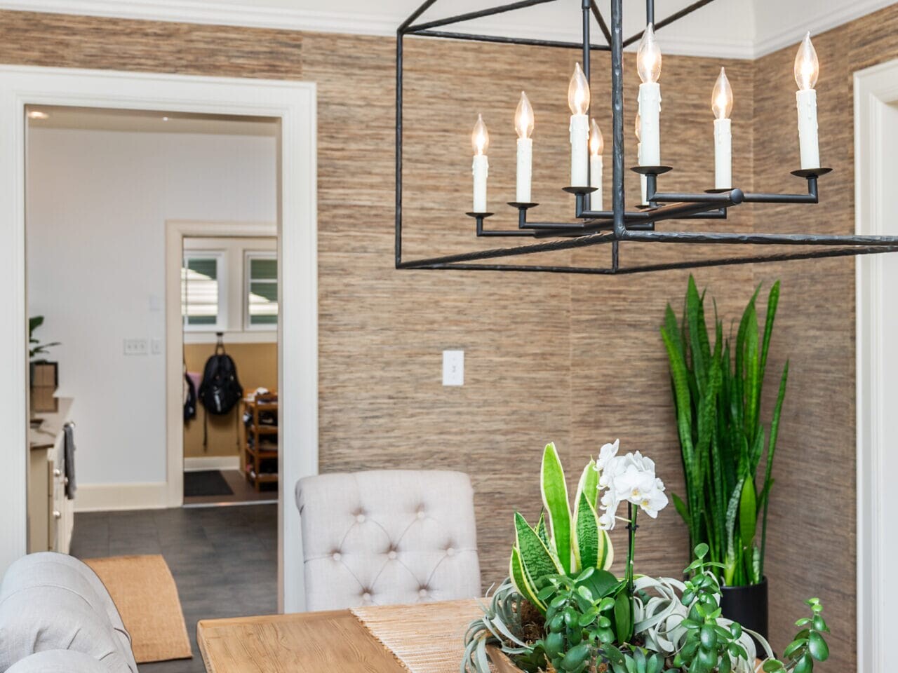 A modern dining room featuring a wooden table with plush beige chairs. A geometric chandelier hangs above, and a centerpiece of green plants decorates the table. In the background, theres a doorway leading to another part of the house.