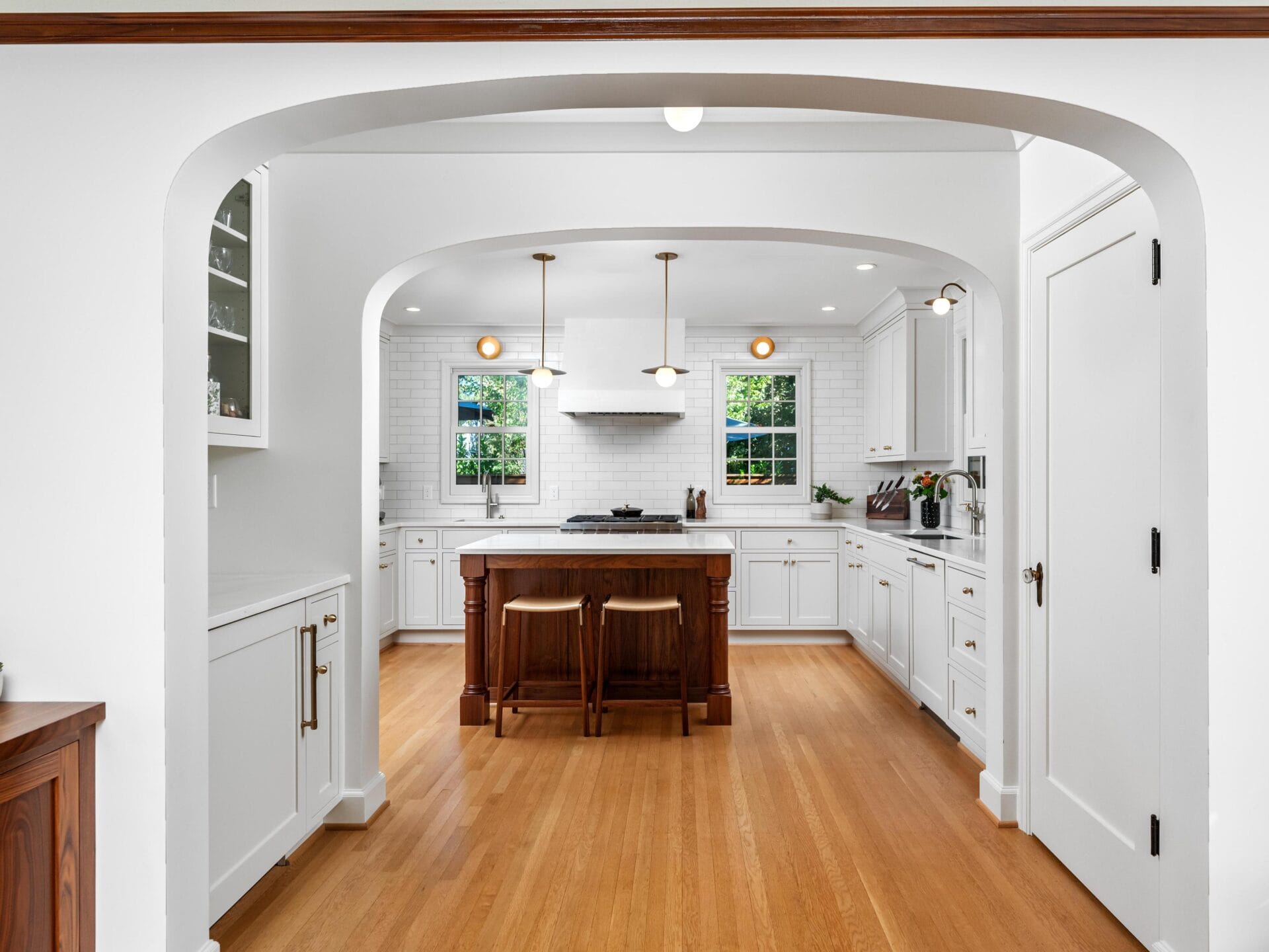 A modern kitchen with white cabinetry, a central island featuring a dark wood base and marble top, two wooden stools, hardwood floors, and two windows bringing in natural light. White subway tiles adorn the walls, and sleek appliances are visible.