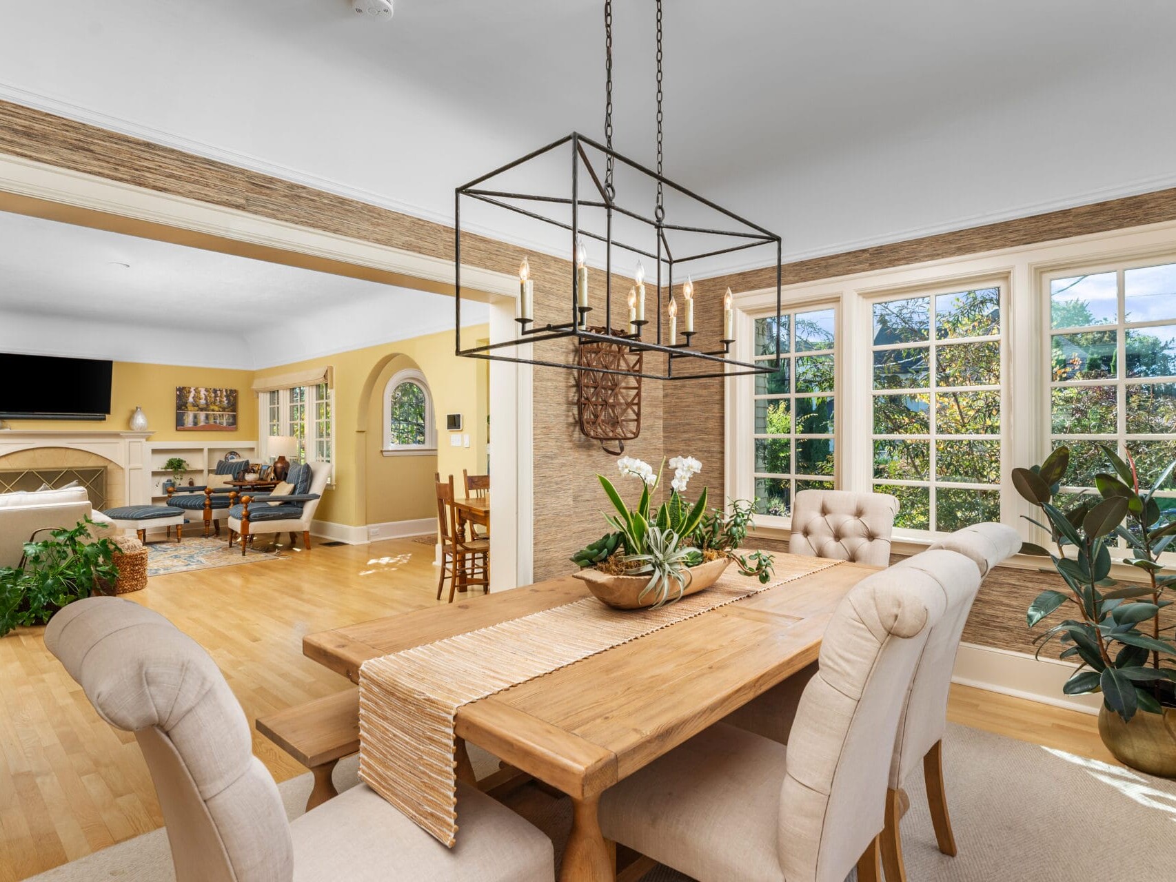 A dining area with a wooden table and cushioned chairs is set in a room with large windows. A geometric chandelier hangs overhead. Adjacent is a living room with a TV, fireplace, and seating. Lush plants decorate the space, enhancing its brightness.