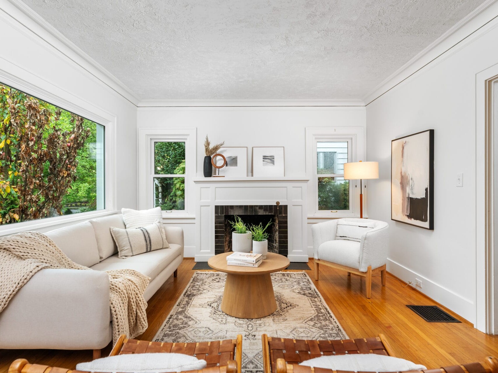 A cozy living room with a white sofa and armchair, a wooden coffee table, and a fireplace. Large windows allow natural light, and art prints decorate the walls. A beige rug covers the wooden floor, creating a warm and inviting atmosphere.