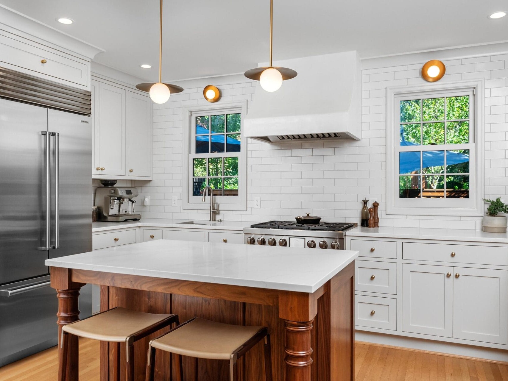 Modern kitchen with white cabinetry, subway tile backsplash, and stainless steel appliances. Wooden island with white countertop and two stools. Two windows provide natural light, and pendant lights hang overhead.