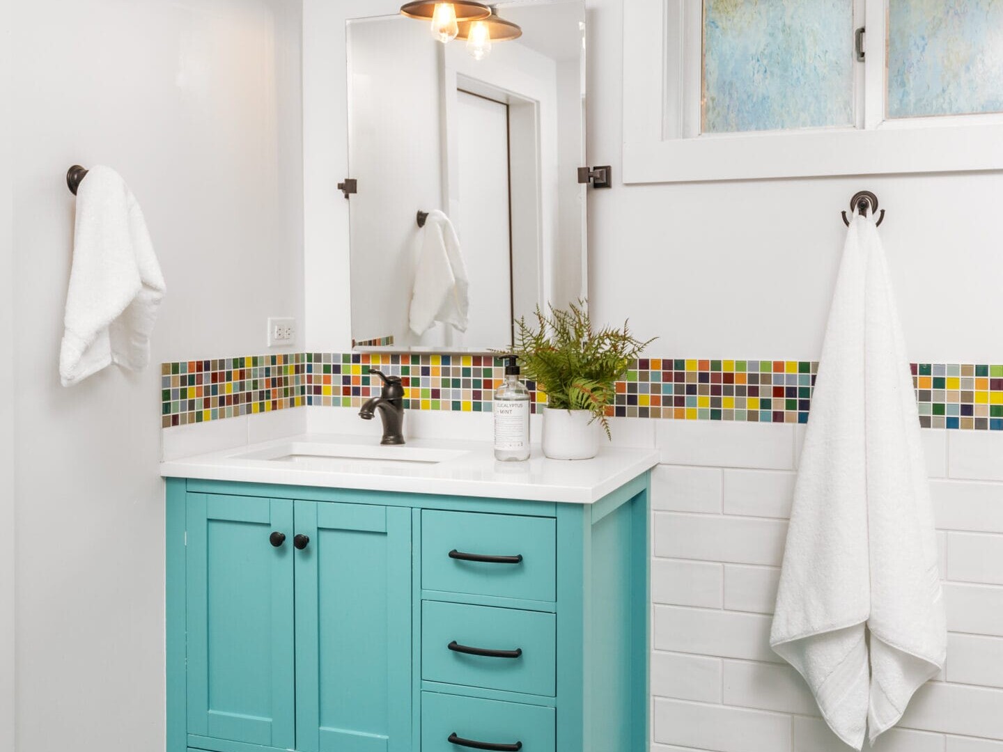 A bathroom with a turquoise vanity, black hardware, and a white countertop. Above is a mirror and light fixture. Walls have colorful mosaic tiles and a white towel hangs on a hook. The floor is striped in blue and teal. A small window is above the sink.