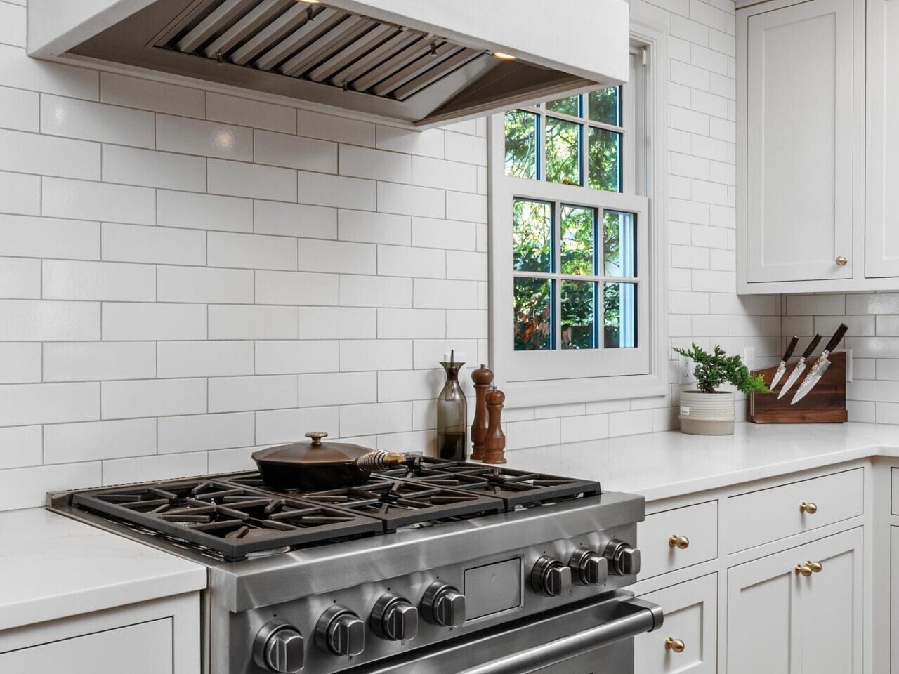A modern kitchen features a stainless steel stove with a white hood against a white subway tile backsplash. Light gray cabinets with gold handles surround the area. A window allows natural light, with cutting boards and a plant placed nearby.