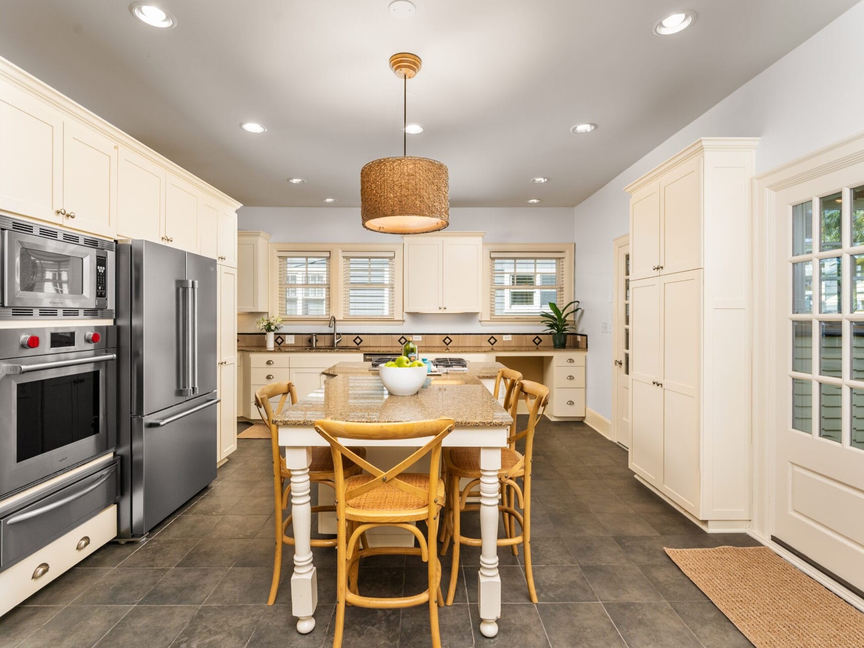 A modern kitchen features white cabinets, stainless steel appliances, and a large island with four wooden chairs. A woven pendant light hangs above the island, and windows allow natural light to fill the space.
