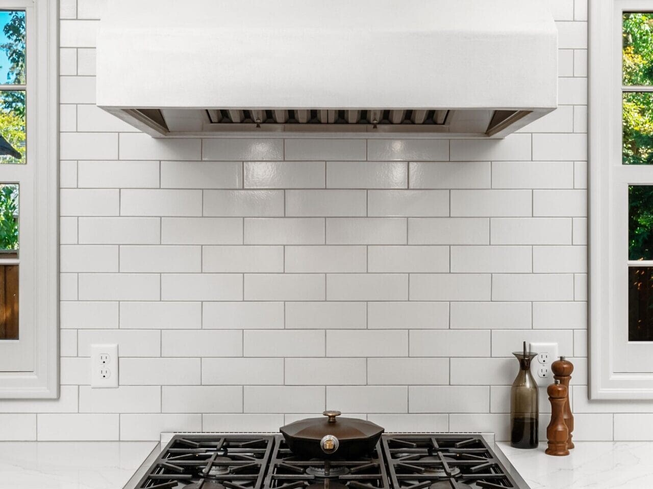 A modern kitchen with a stainless steel stove featuring a pot on the burners. Above is a large white vent hood. The backsplash is made of white subway tiles, and there are windows on either side letting in natural light.