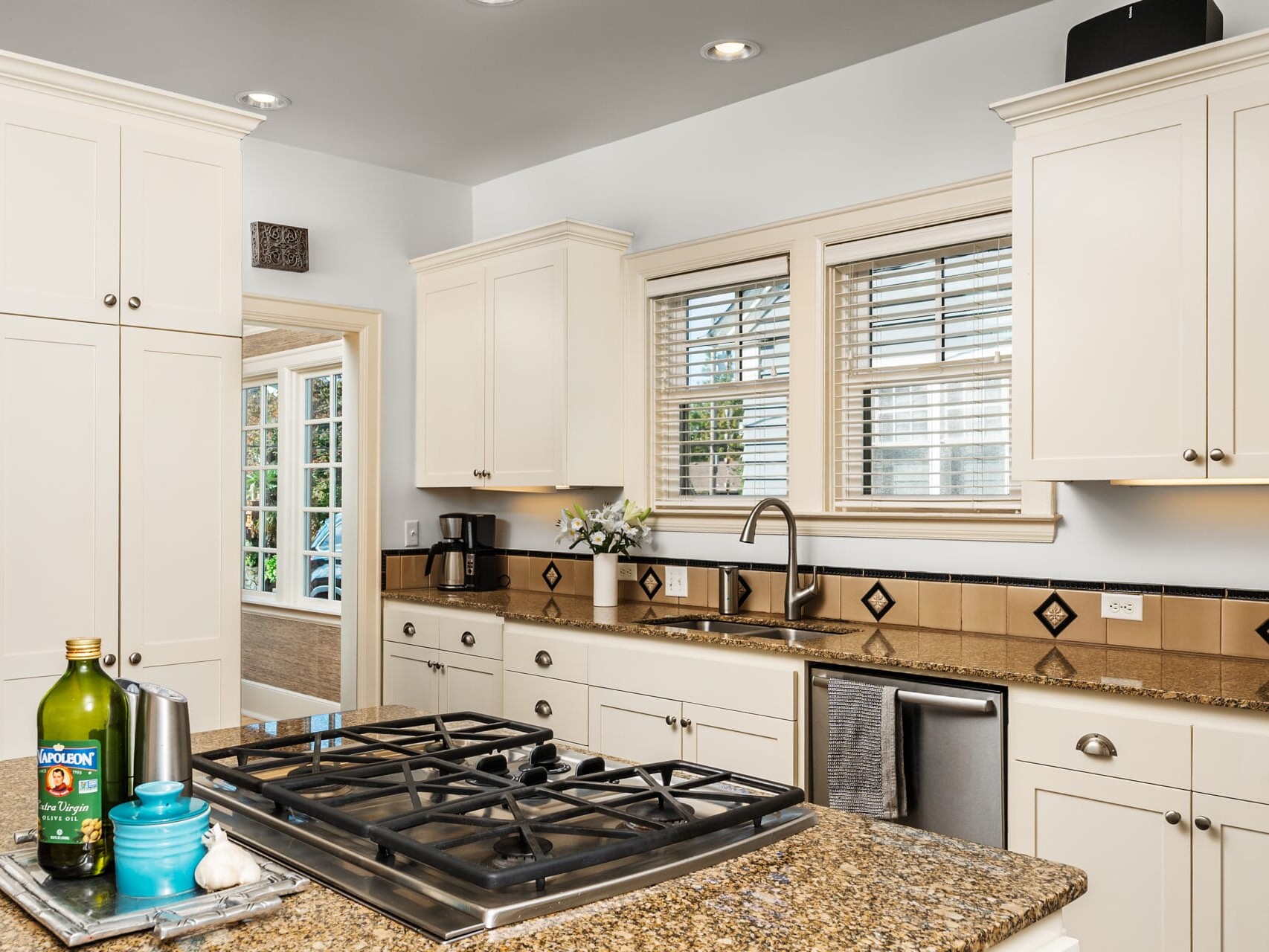 A kitchen with white cabinets, granite countertops, and a built-in gas stovetop. A bottle of olive oil and a blue spice container are on the counter. A window above the sink lets in natural light, and the doorway leads to a bright adjoining room.