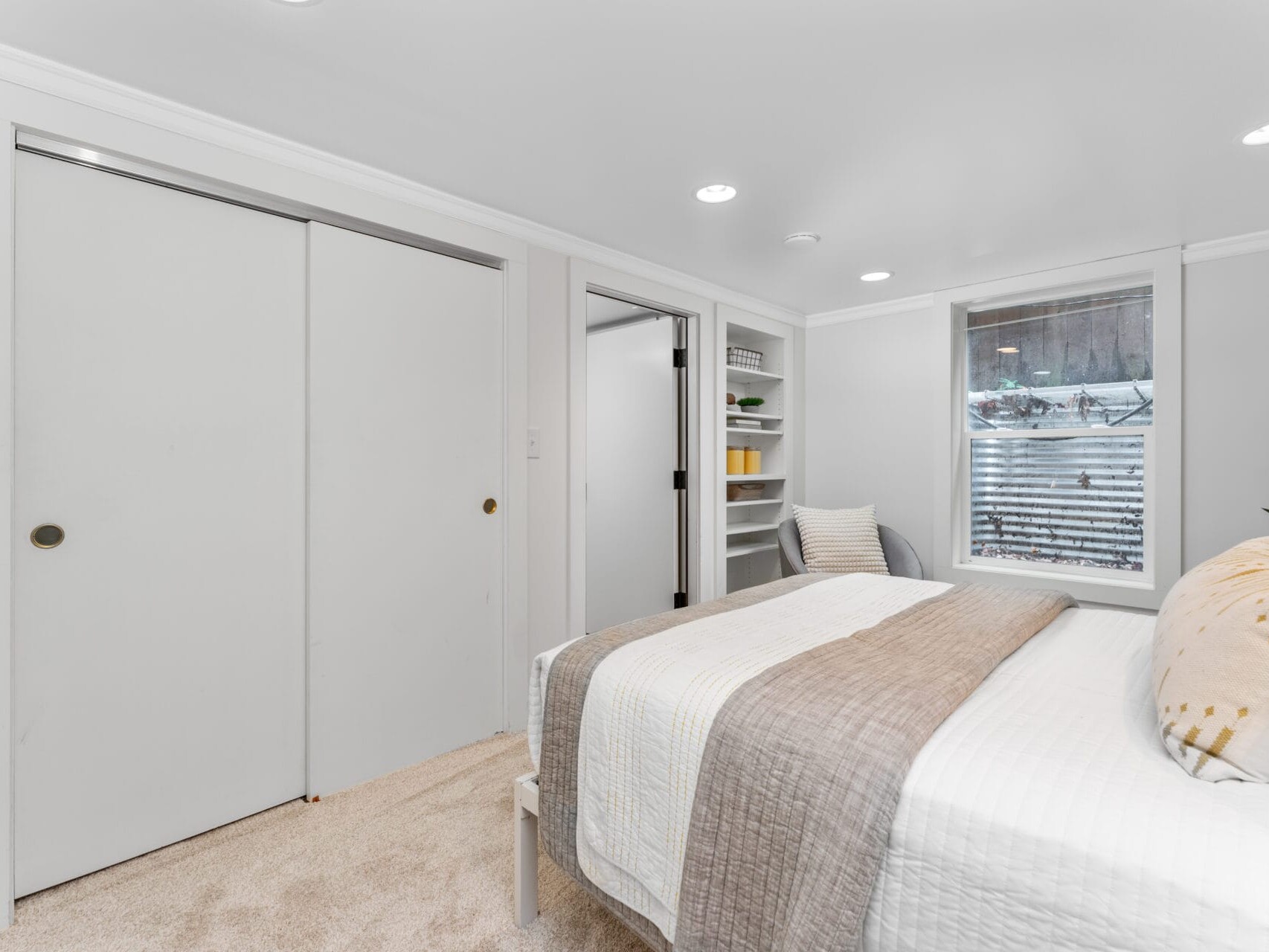 A minimalist bedroom with white walls and beige carpet features a bed with white and beige linens. There are sliding closet doors, a window with blinds, and built-in shelves with colorful decor. A small plant adds a touch of greenery.