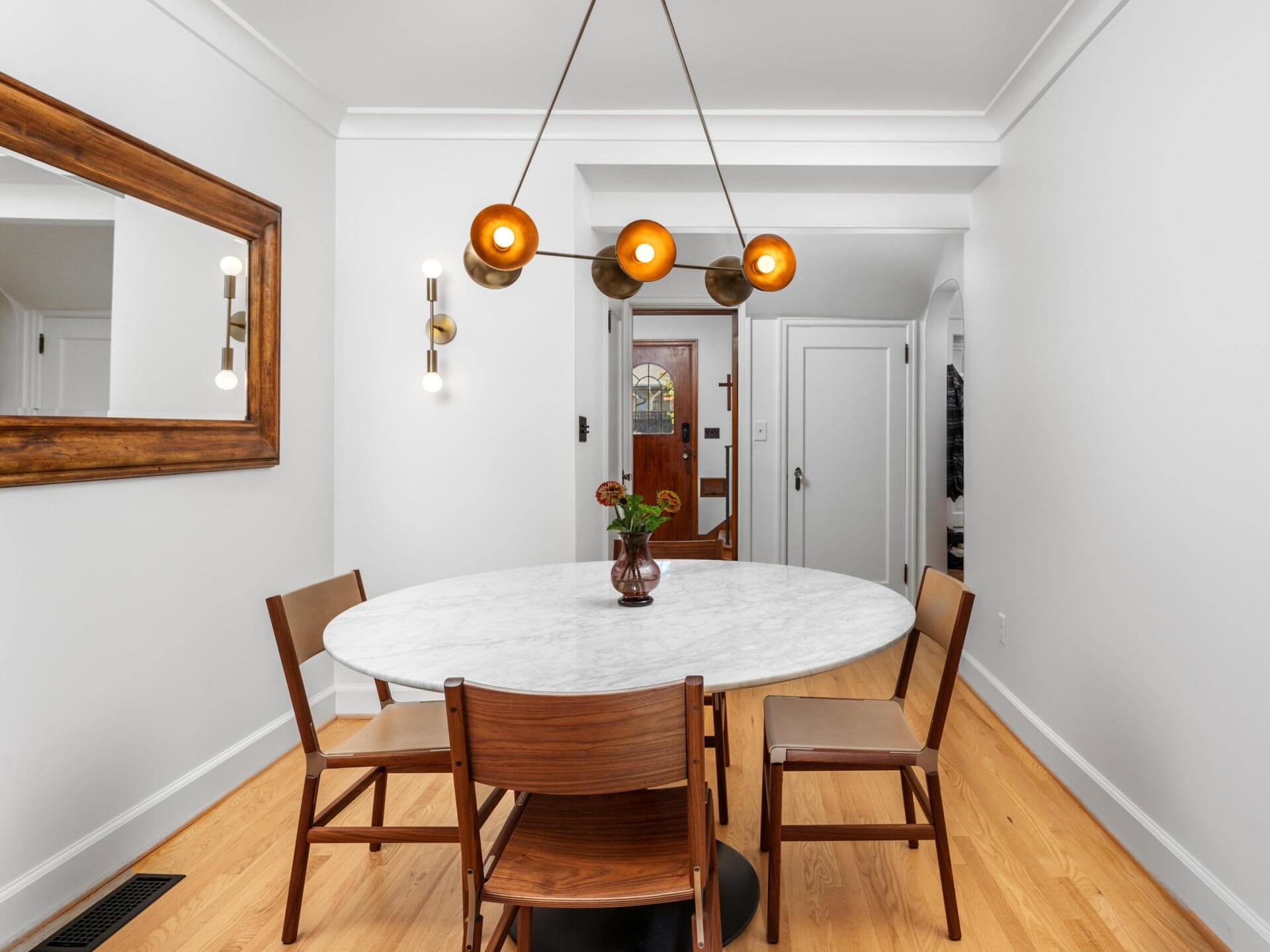 A modern dining room with a round marble table and three wooden chairs. A unique light fixture with glowing bulbs hangs above. The room features white walls, a large wooden mirror, and hardwood flooring. A vase with flowers sits on the table.
