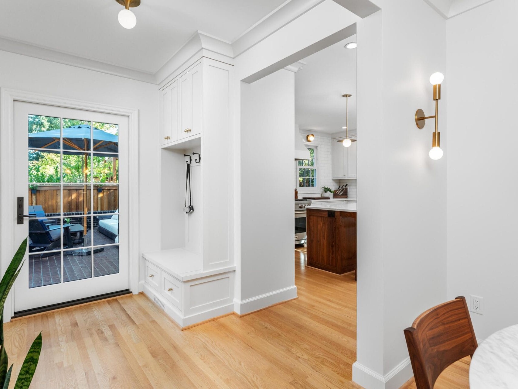 A bright and airy interior featuring a white kitchen with wooden floors. A dining area is partially visible, and a door with glass panels leads to a patio with outdoor furniture. Natural light enters through the windows, highlighting the modern decor.