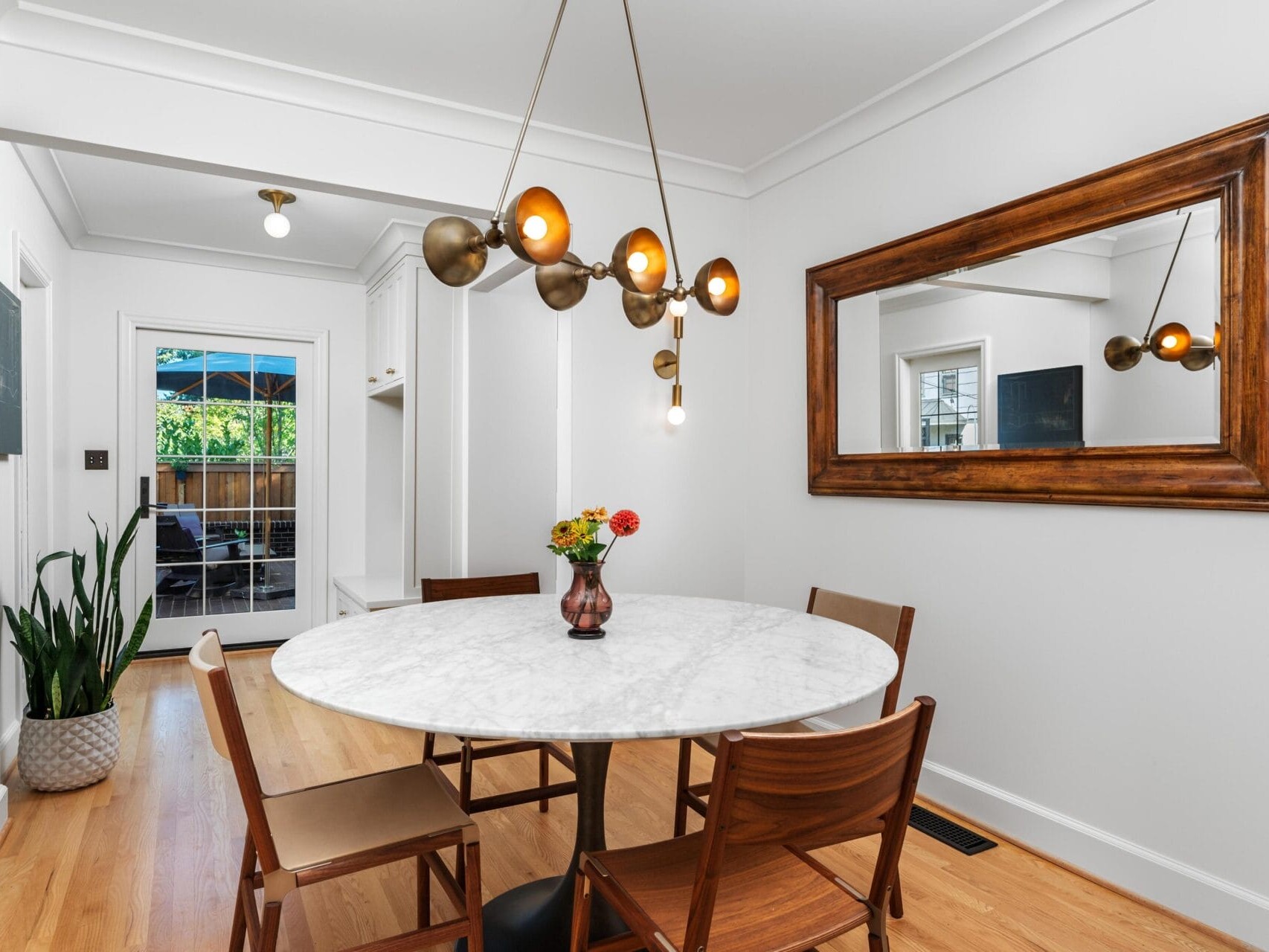 A modern dining room features a round marble table surrounded by wooden chairs. A contemporary chandelier hangs above, and a large mirror is mounted on the wall. Potted plants and a glass door leading outside complete the setting.