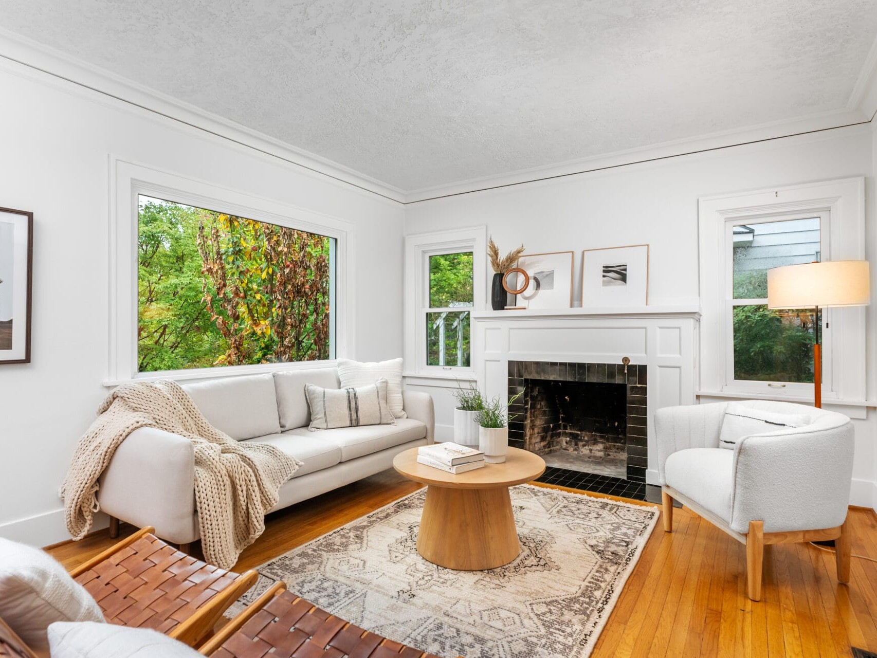 A cozy living room with a white sofa, armchair, and wooden chairs arranged around a round wooden coffee table. A fireplace is flanked by windows and decorated with minimalist decor. Large window shows greenery outside. Soft lighting from a floor lamp.