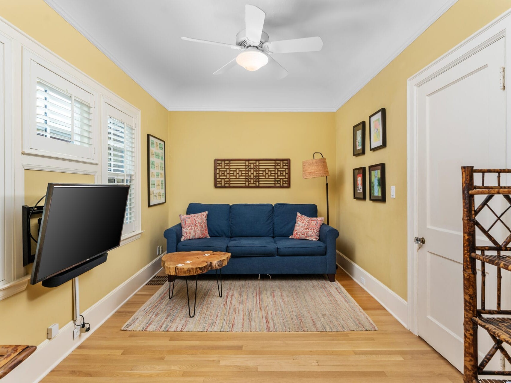 A cozy living room with yellow walls, featuring a blue sofa with decorative pillows. A wooden coffee table sits on a rug. Artwork adorns the walls. A ceiling fan is above, and a flat-screen TV is mounted on the side wall. Natural light filters through the windows.