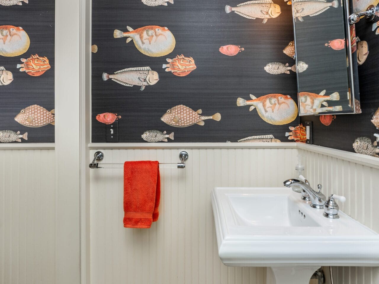 A small bathroom with fish-patterned wallpaper and cream wainscoting. Features include a white pedestal sink, mirror with overhead lights, a red towel on the towel rack, and a beige woven mat on the tile floor.