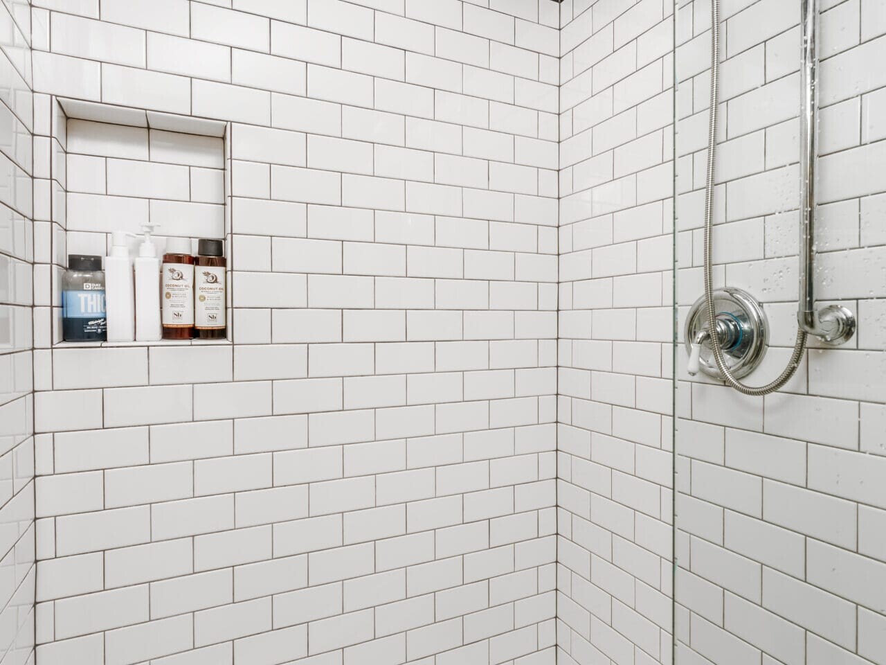 A modern shower features white subway tiles and a built-in shelf holding toiletries. The ceiling trim is decorated with colorful fish patterns. A silver showerhead and glass partition complete the look. The floor has white hexagonal tiles.
