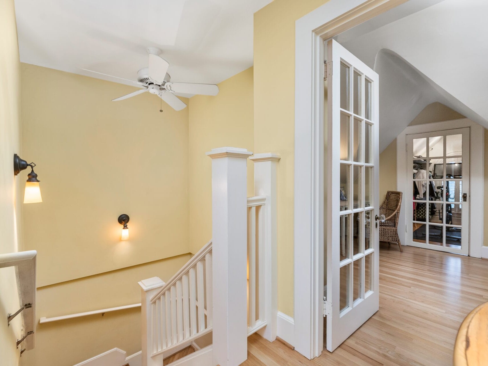 A bright staircase area with light yellow walls, white trim, and wooden floors. A ceiling fan and black wall lamps illuminate the space. French doors open to a room with a sloped ceiling and wooden furniture.
