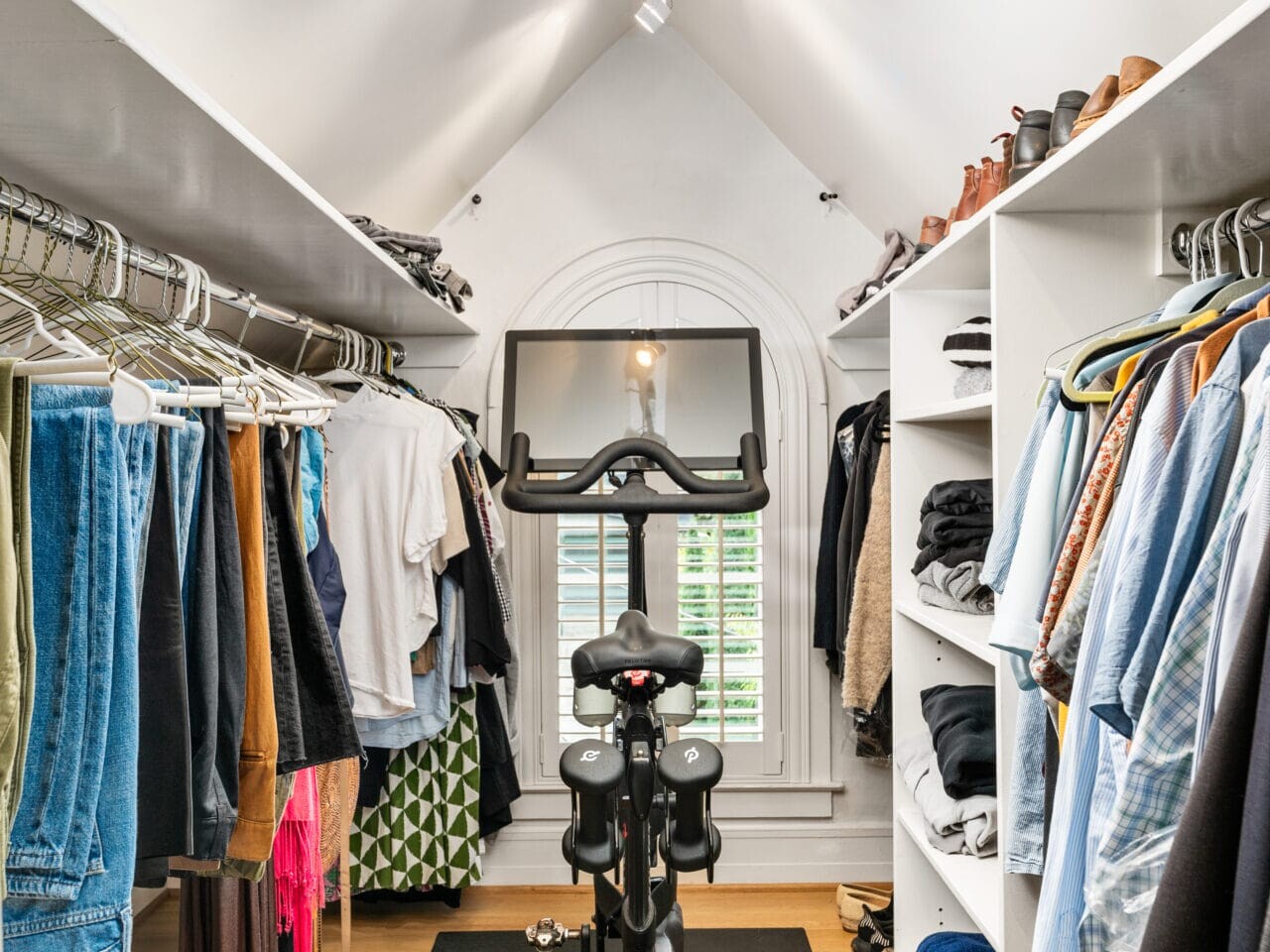 A small, well-organized walk-in closet with clothes hanging on both sides and folded on shelves. A stationary exercise bike is positioned on a black mat in the center under a vaulted ceiling with track lighting.