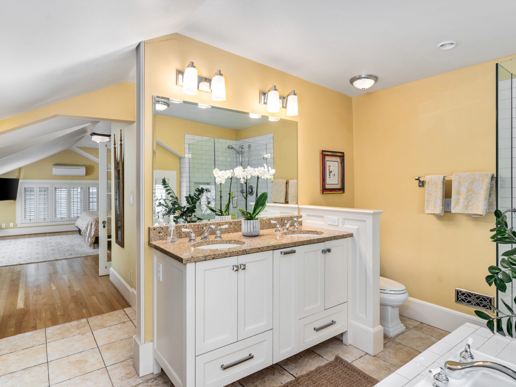 A spacious bathroom with yellow walls features a granite countertop vanity and a large mirror, lit by wall sconces. A shower with glass doors is visible. Decorative plants and flowers add a touch of greenery. The room leads to a bedroom with wooden flooring.