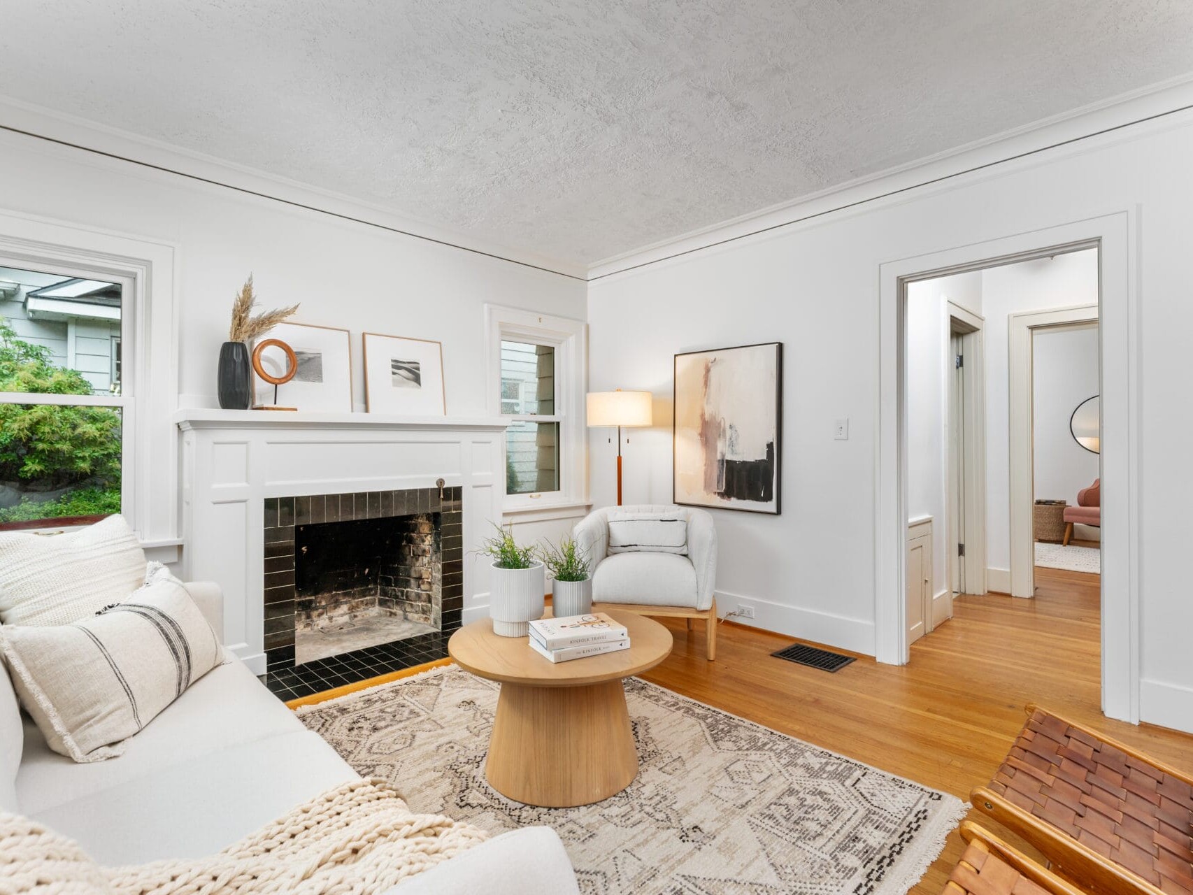 A cozy living room with a white and beige color scheme. It features a white sofa, a wooden coffee table, a fireplace with decorative items, and a lamp. Wooden flooring and two chairs complete the decor. A hallway leads to another room.