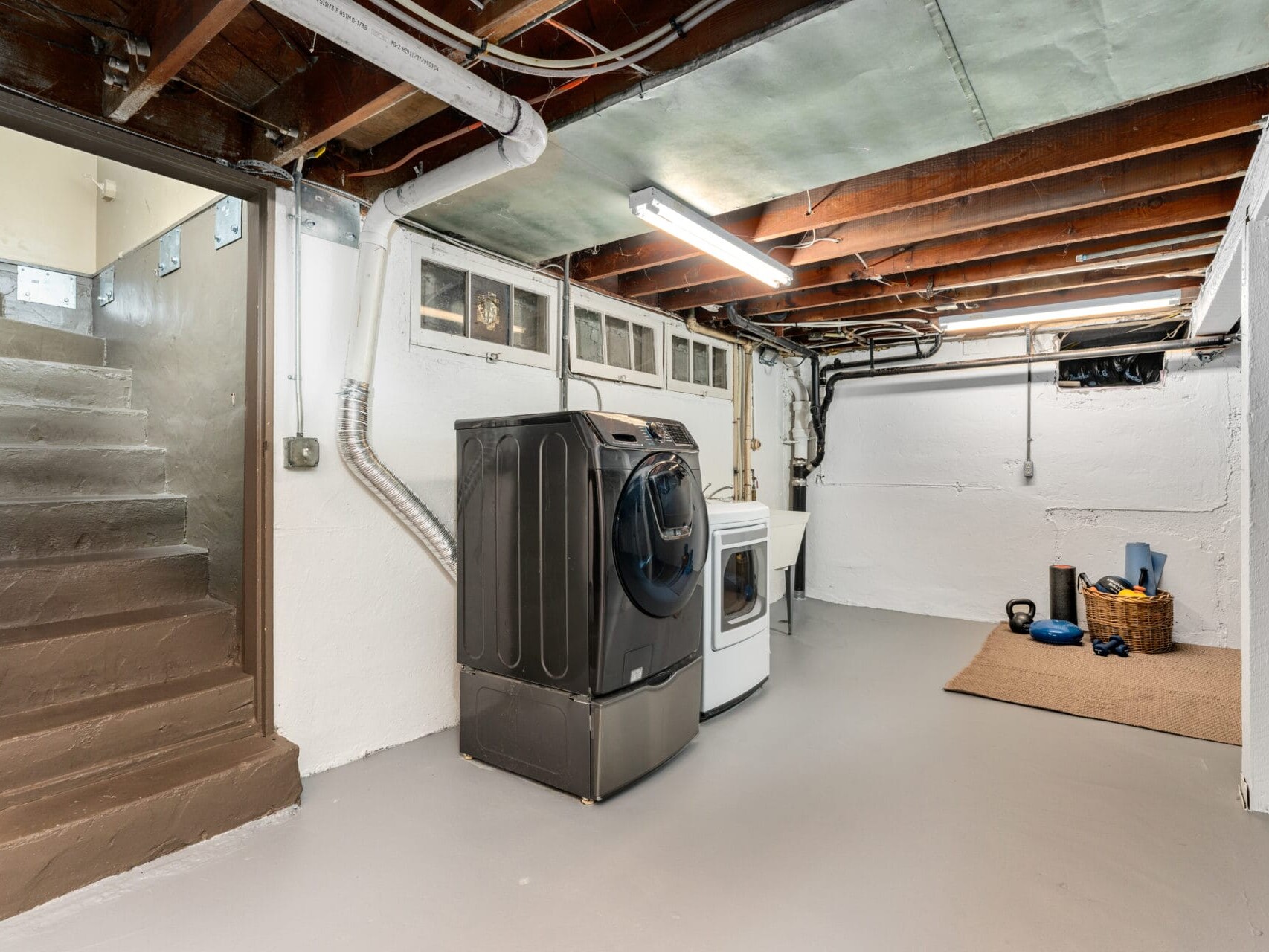 A basement laundry room with exposed beams, featuring a black front-loading washing machine and a white dryer. The space includes a yoga mat, a basket with towels, and some fitness equipment on the floor. Stairs lead up to the next level.