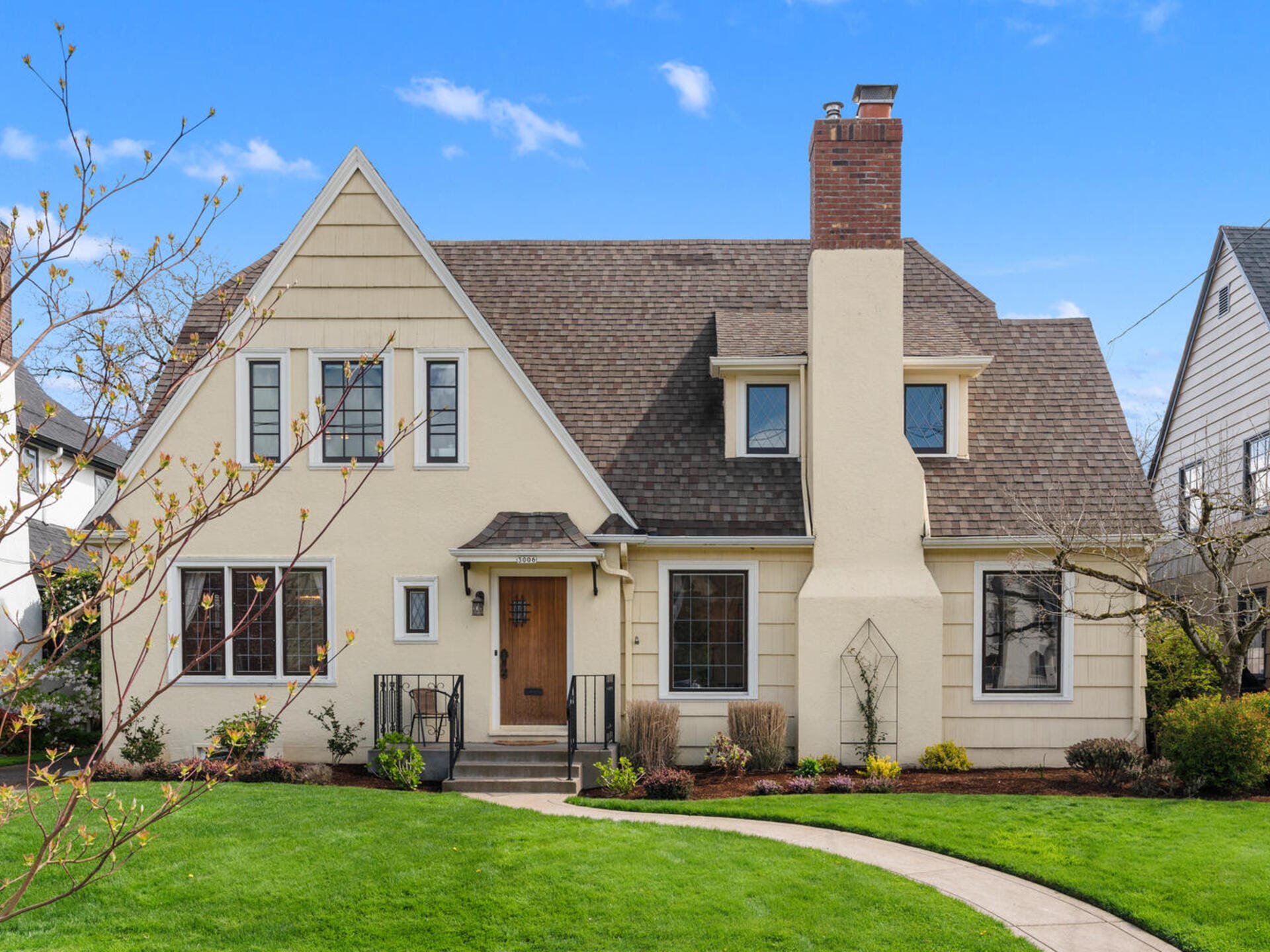 A charming two-story, beige house with a steeply pitched roof and brick chimney. It features a front door with a small porch, surrounded by windows. A neatly manicured lawn and pathway lead to the entrance, with a clear blue sky in the background.