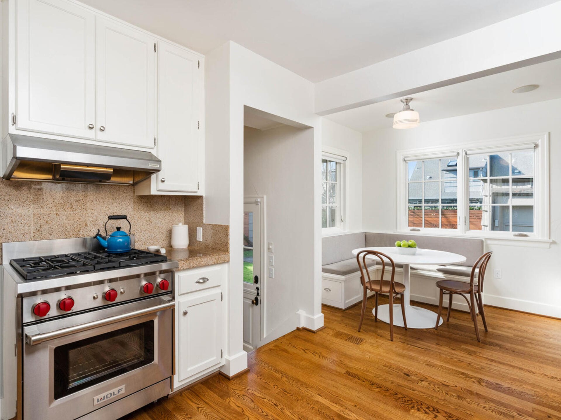 A bright kitchen with hardwood floors features a stove, oven, and cabinetry on the left. A cozy breakfast nook with a round table and chairs is in front of two windows. A blue kettle sits on the stove, and a fruit bowl is on the table.