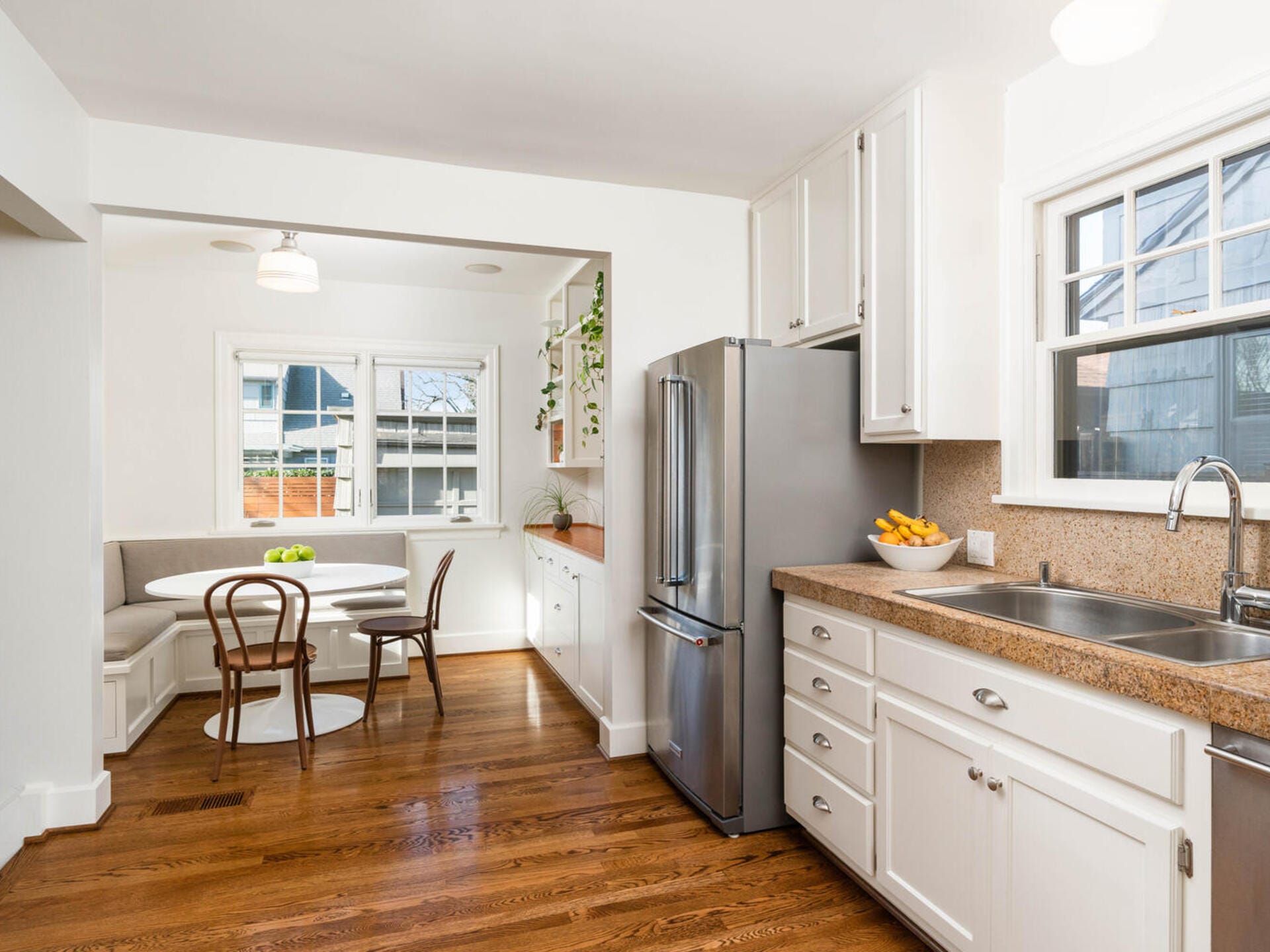 A bright kitchen with wooden floors, white cabinets, and a stainless steel refrigerator. A round table with chairs sits in a cozy nook with a cushioned bench by the window. The countertop has a bowl of bananas and green apples. A small plant decorates a shelf.
