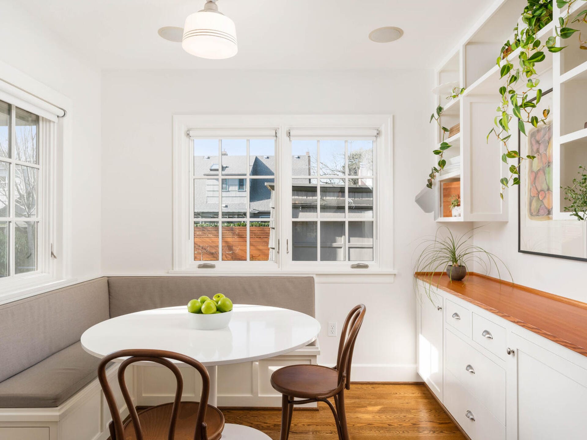 A bright, cozy dining nook with a white table, two wooden chairs, and a built-in bench with gray cushions. Windows let in natural light, and there are plants on shelves and a bowl of green apples on the table.