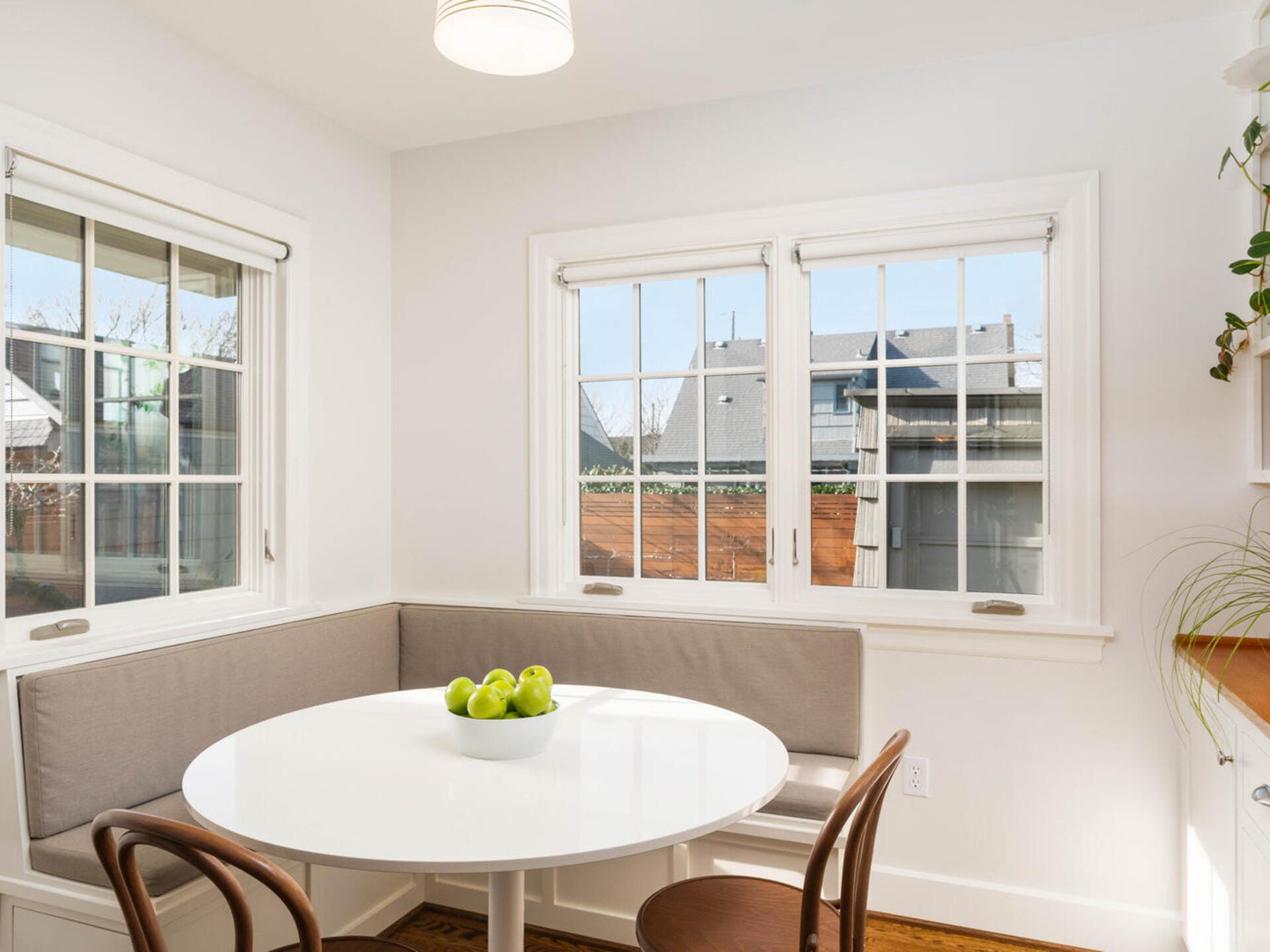 A bright, cozy dining nook with a white, round table and gray-cushioned benches. Wooden chairs surround the table, and a bowl of green apples is in the center. Large windows provide natural light and an outside view. A hanging light fixture is above.
