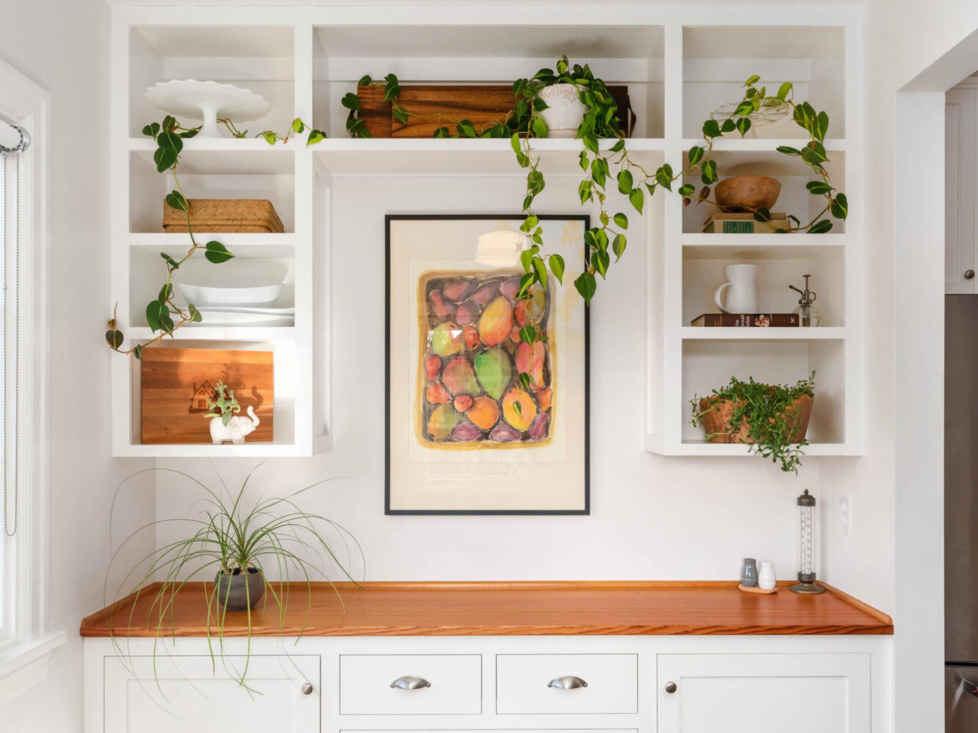 A white kitchen with wooden countertops features open shelves filled with plants and dishes. A colorful fruit art print is centered on the back wall. Minimalistic decor includes a plant cascading from the top shelf and bright natural lighting.