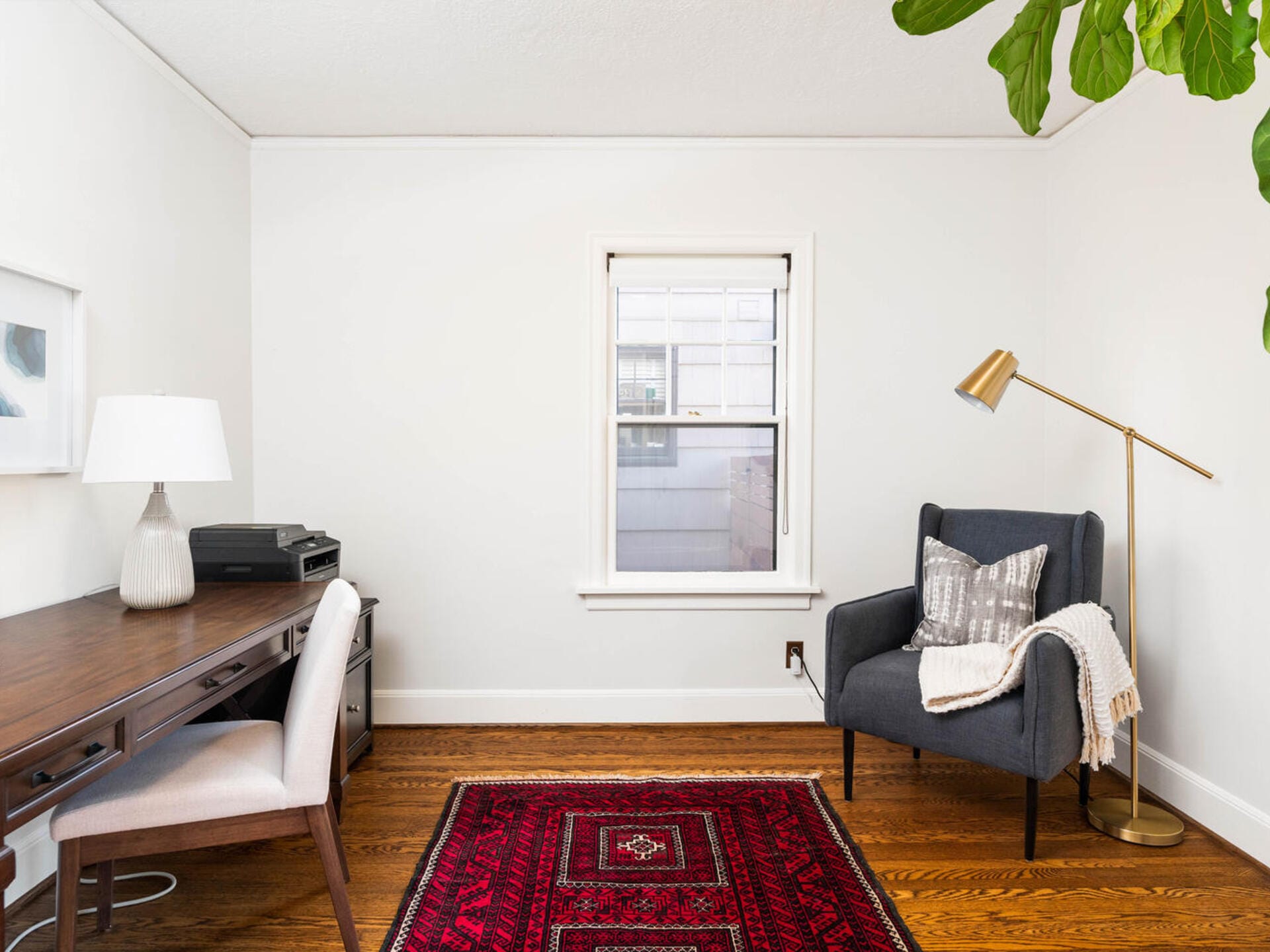 A cozy home office with a wooden desk, chair, lamp, and printer on the left. To the right, a dark armchair with a blanket sits under a brass floor lamp. A red patterned rug is on the wooden floor, and a window is centered on the white wall.