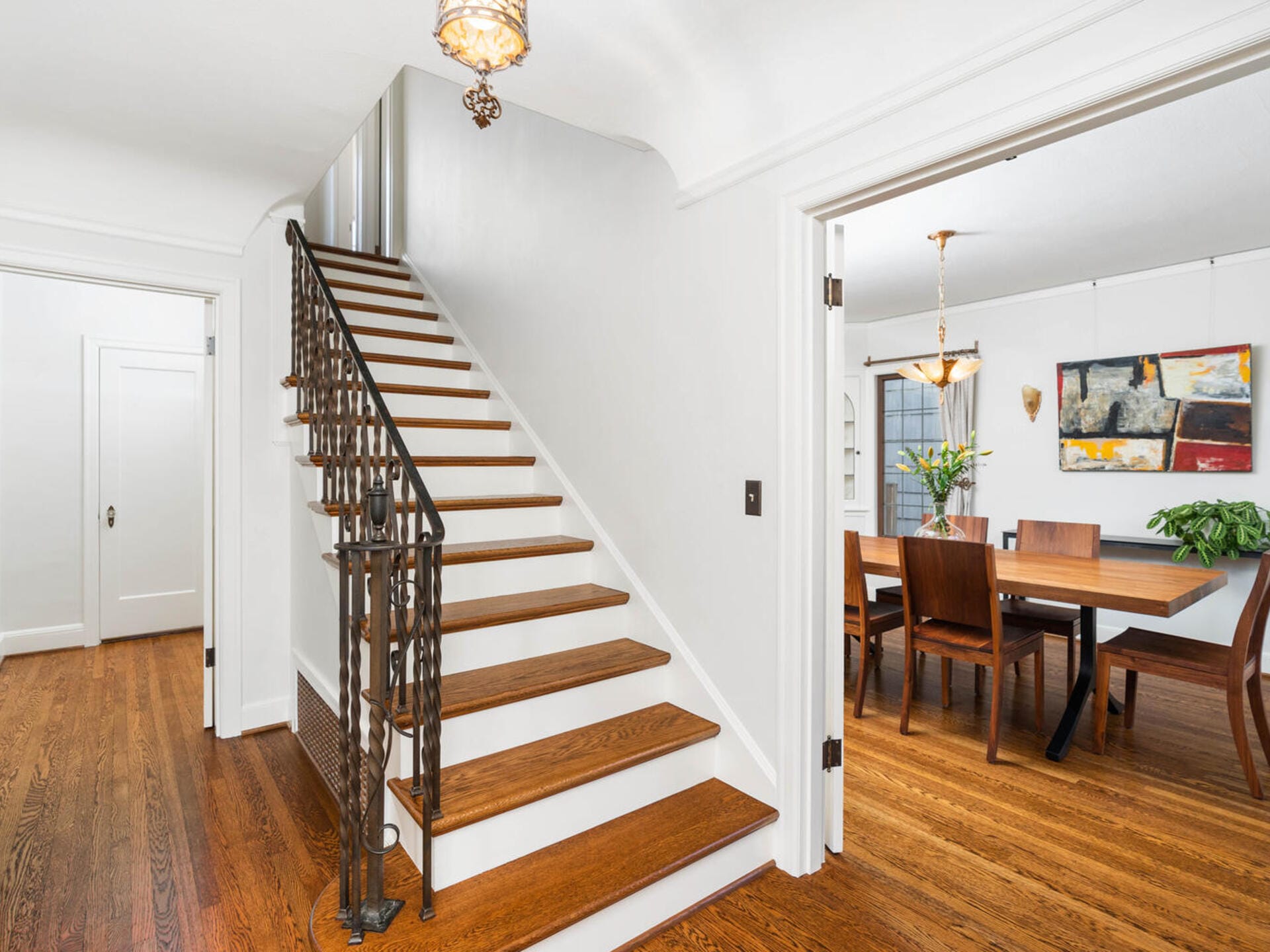 A bright dining room with a wooden table and chairs is visible through a doorway. Adjacent is a staircase with wooden steps and an ornate black railing. The floors are hardwood, and a decorative light fixture hangs from the ceiling.