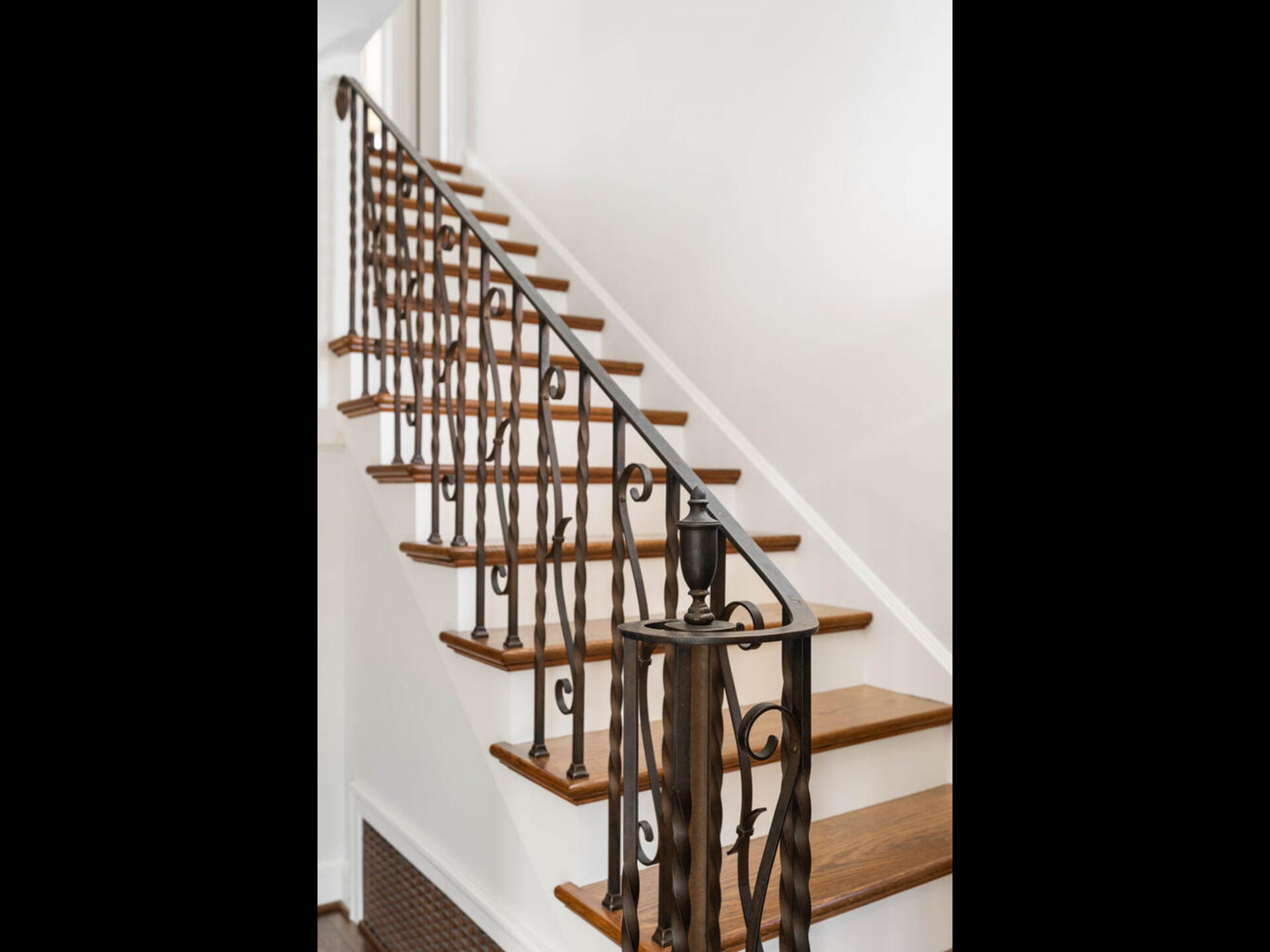 Wooden staircase with dark ornate iron railing, leading up alongside a white wall. The steps feature dark wood treads and white risers, creating a classic, elegant look.