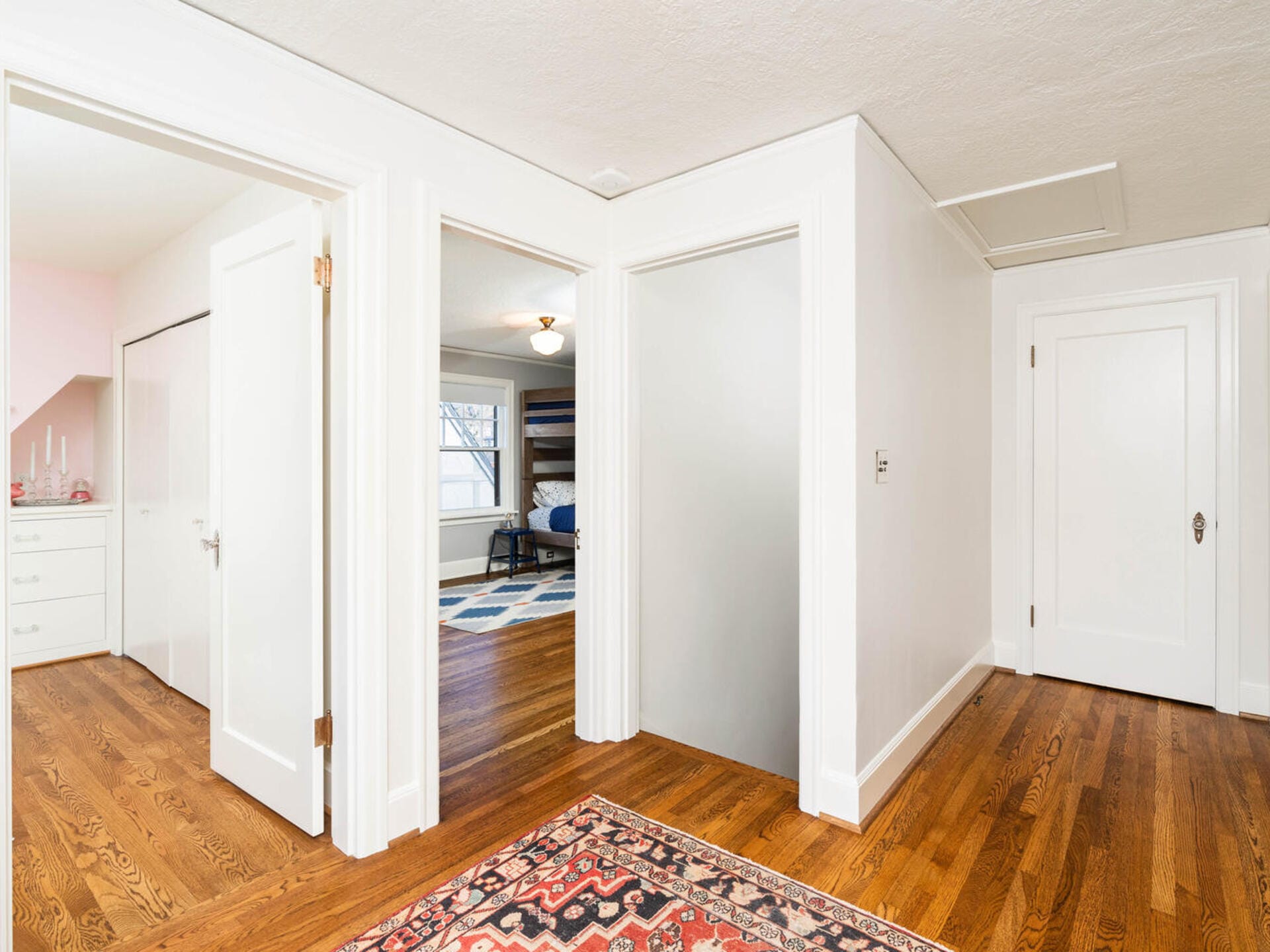 A bright hallway with wooden floors and white walls. Three doors open into different rooms, including a bedroom with bunk beds and a walk-in closet with drawers. A patterned rug lies on the floor, and a window offers natural light.